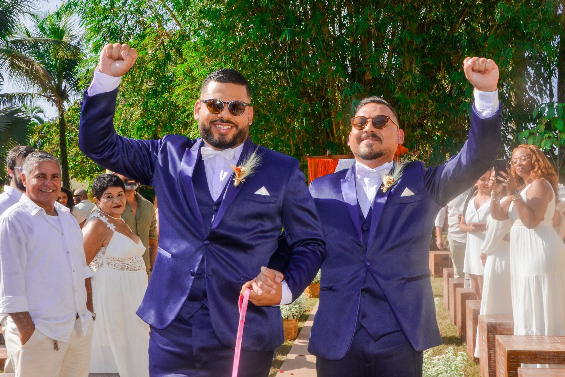 Dois homens de terno azul levantam os punhos, comemorando seu casamento ao ar livre.