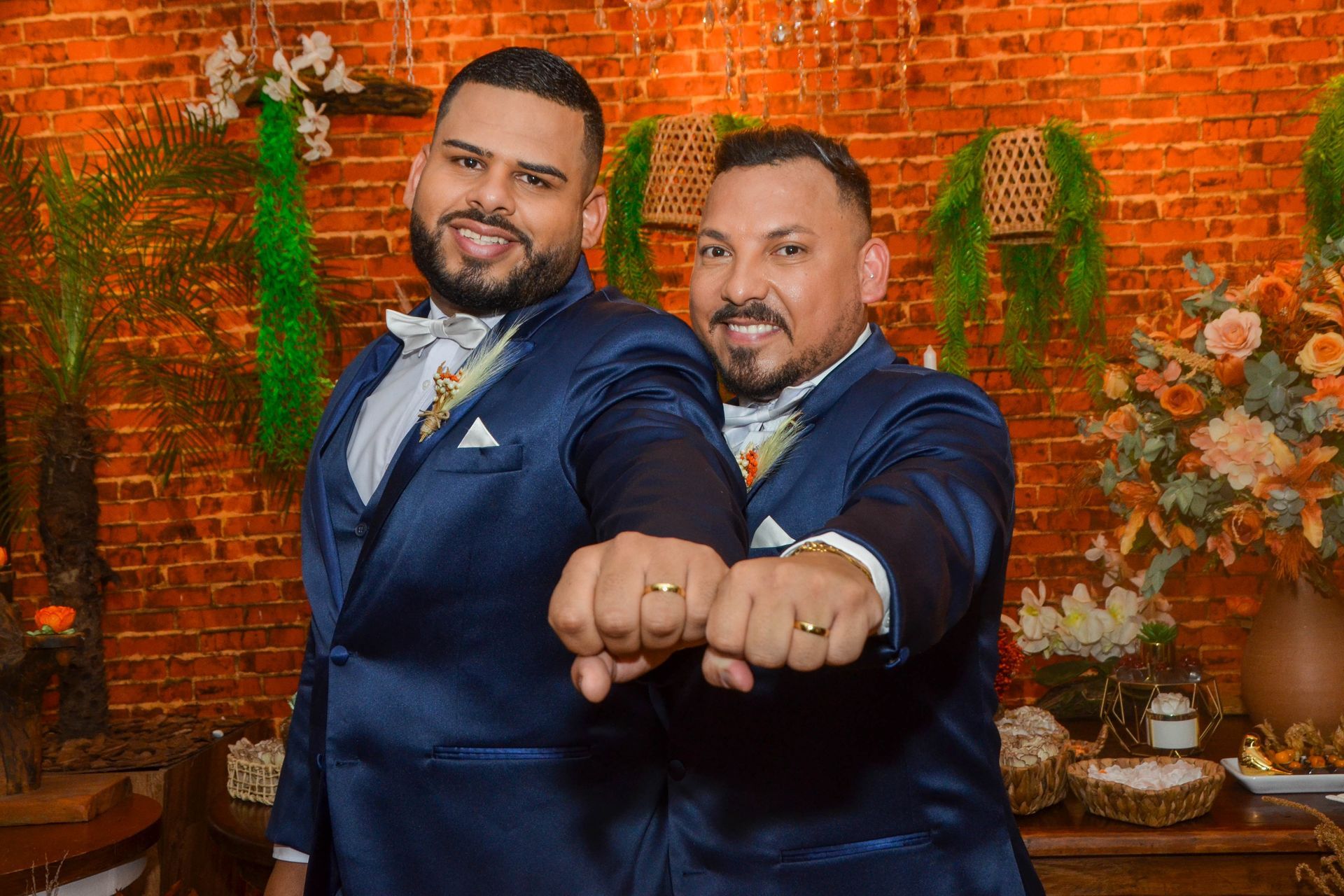 Dois homens de terno azul exibem alianças, sorrindo para a câmera, em frente a uma parede de tijolos laranja.