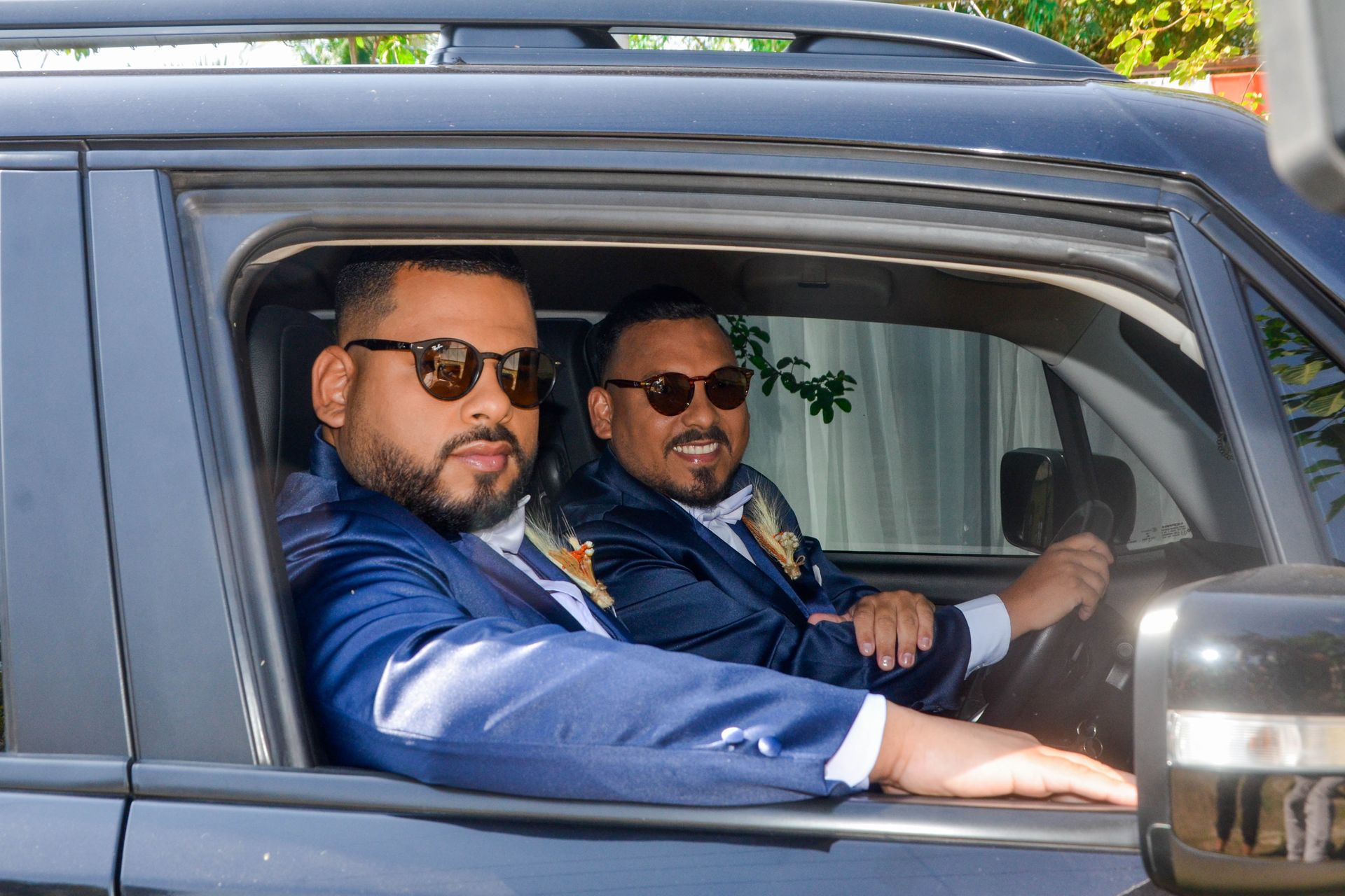 Dois homens de terno azul e óculos de sol em um carro preto, sorrindo.