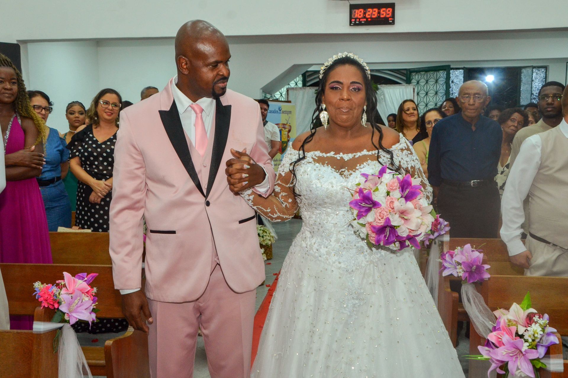 Casal caminhando pelo corredor após o casamento; noiva em vestido branco, noivo em terno rosa; igreja.