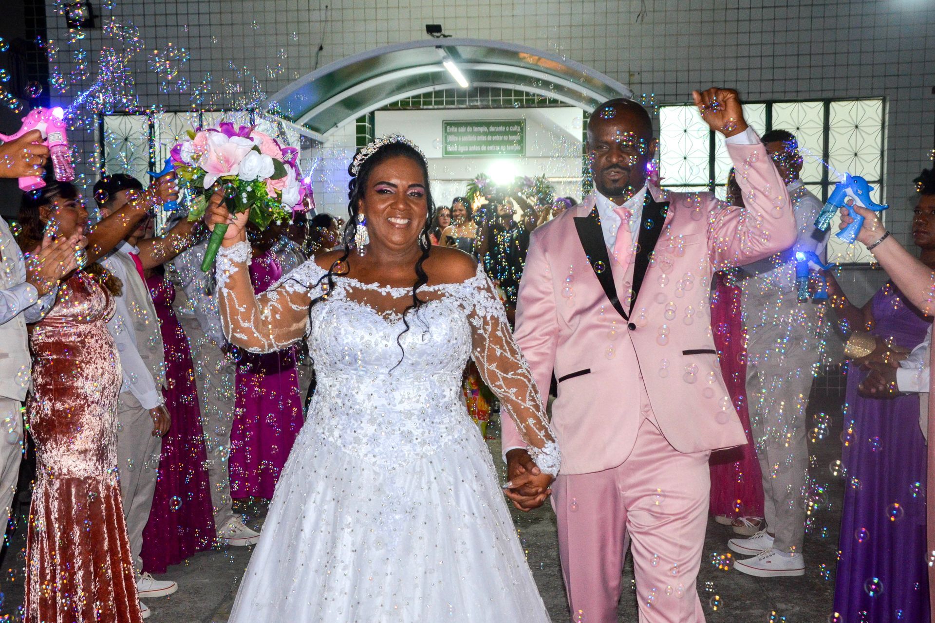 Recém-casados saindo do prédio, de mãos dadas, braços levantados; confete e bolhas os cercam.
