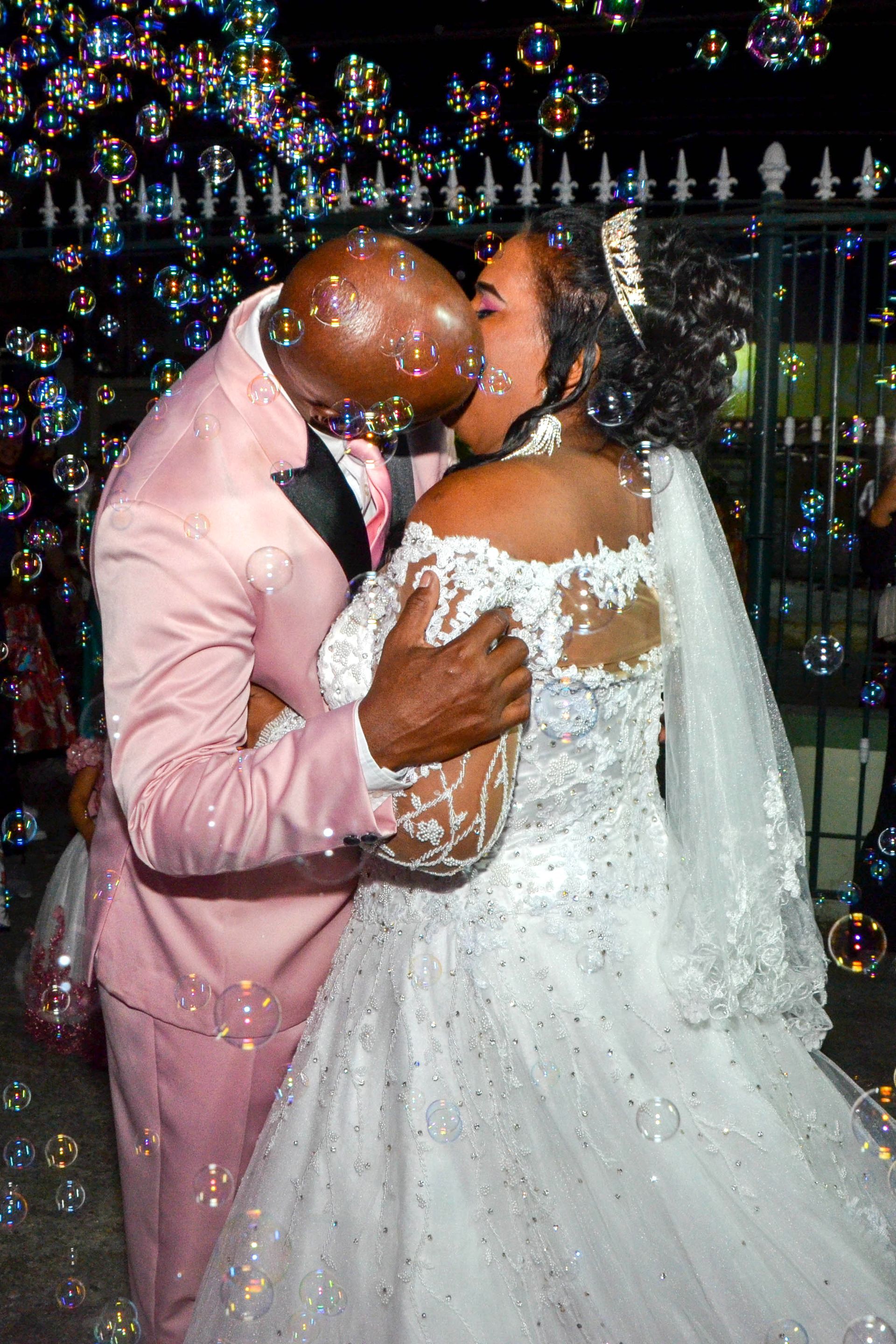 Casal se beijando, cercado por bolhas, a noiva em um vestido de noiva branco, o noivo em um terno rosa.