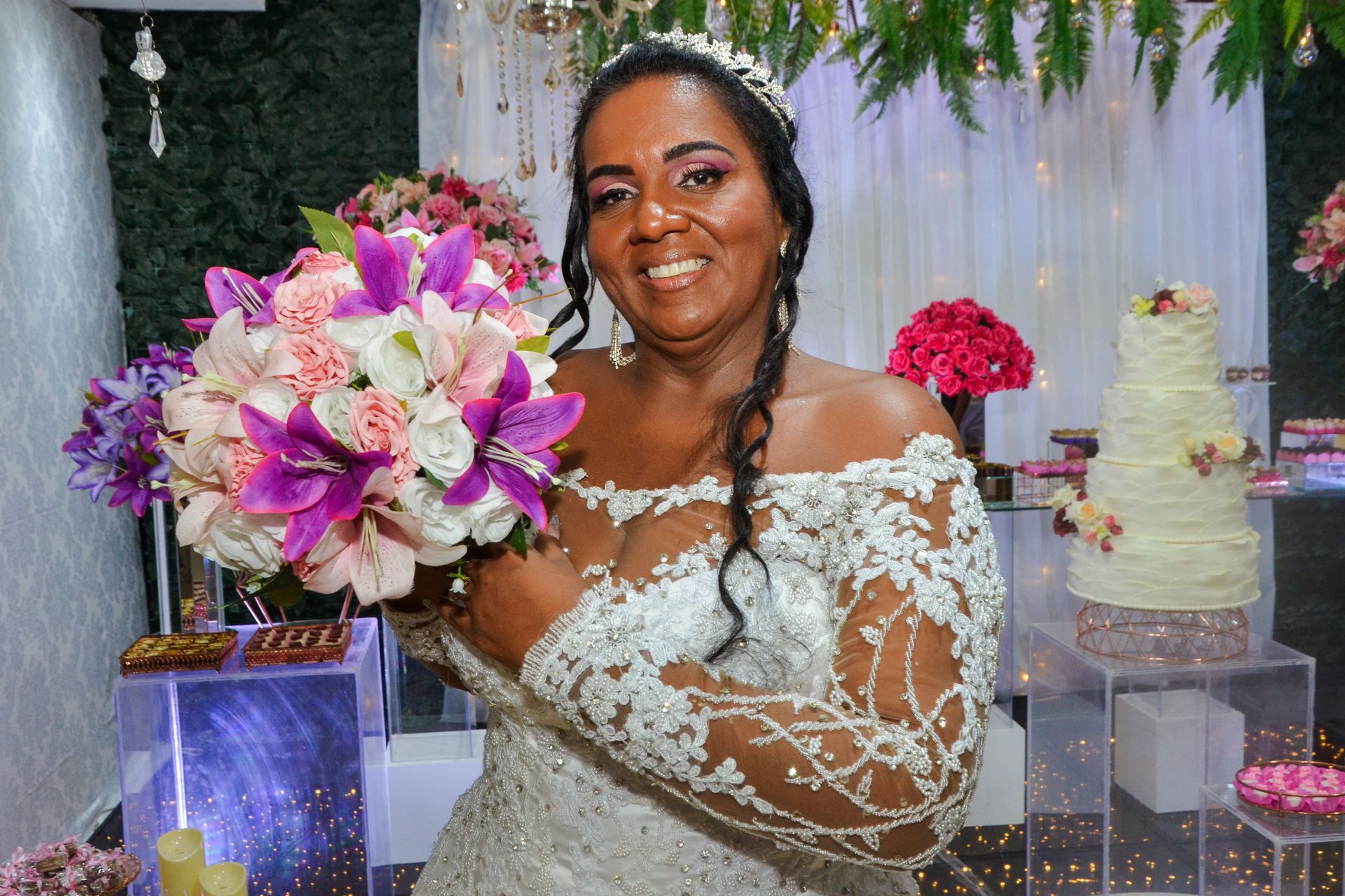 Noiva em vestido de renda segurando um buquê, sorri perto de um bolo de vários andares.