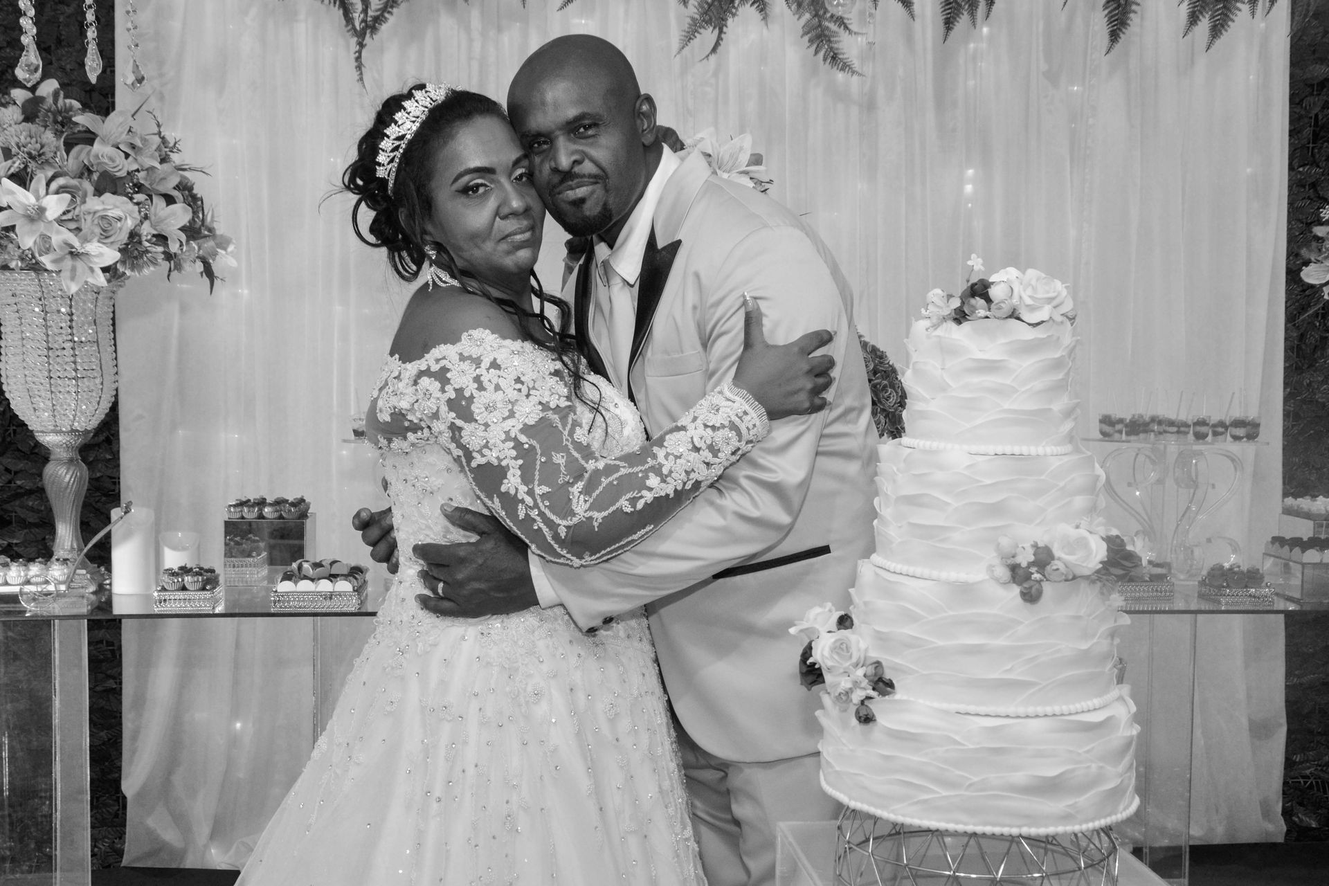 Noivos se abraçam em frente a um bolo de casamento de vários andares. A noiva usa um vestido de renda. O noivo usa um terno leve.
