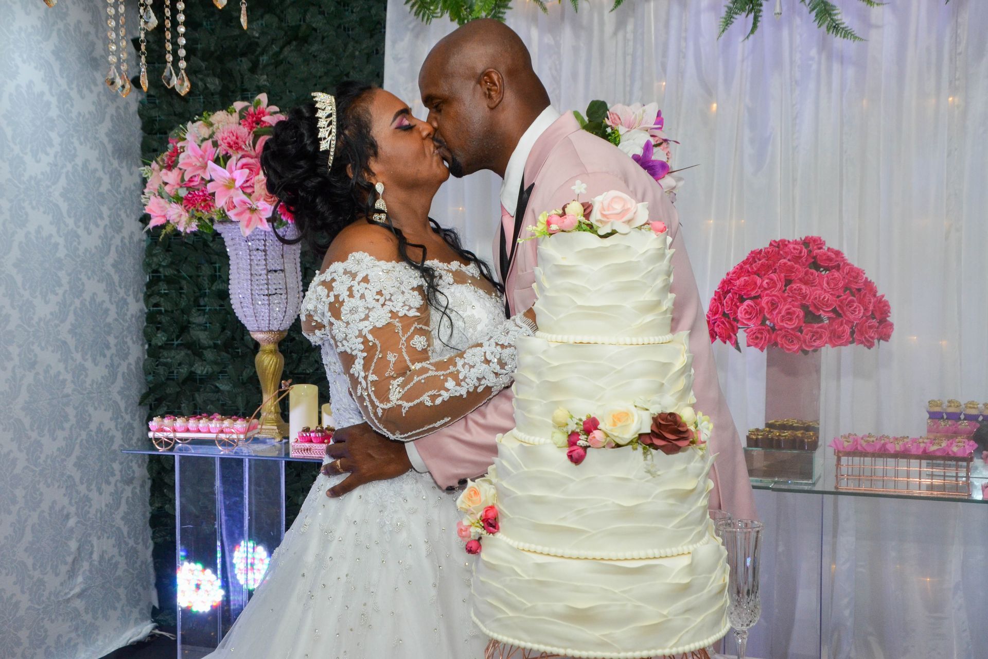 Casal se beija ao lado do bolo de casamento na recepção, decorado com flores e detalhes em rosa.