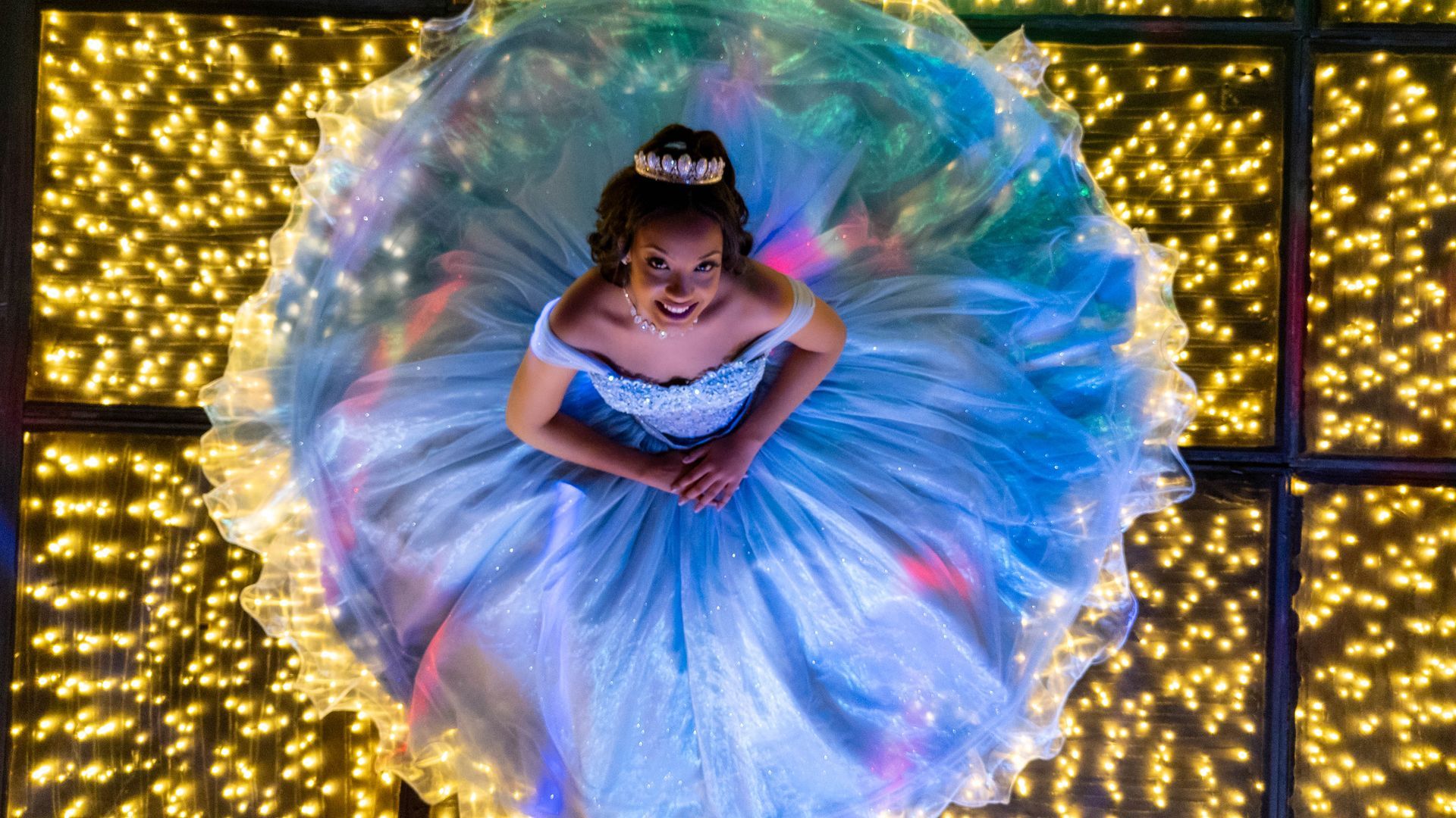Garota de vestido azul com tiara, olhando para cima, em pé sobre um chão iluminado por luzes amarelas.