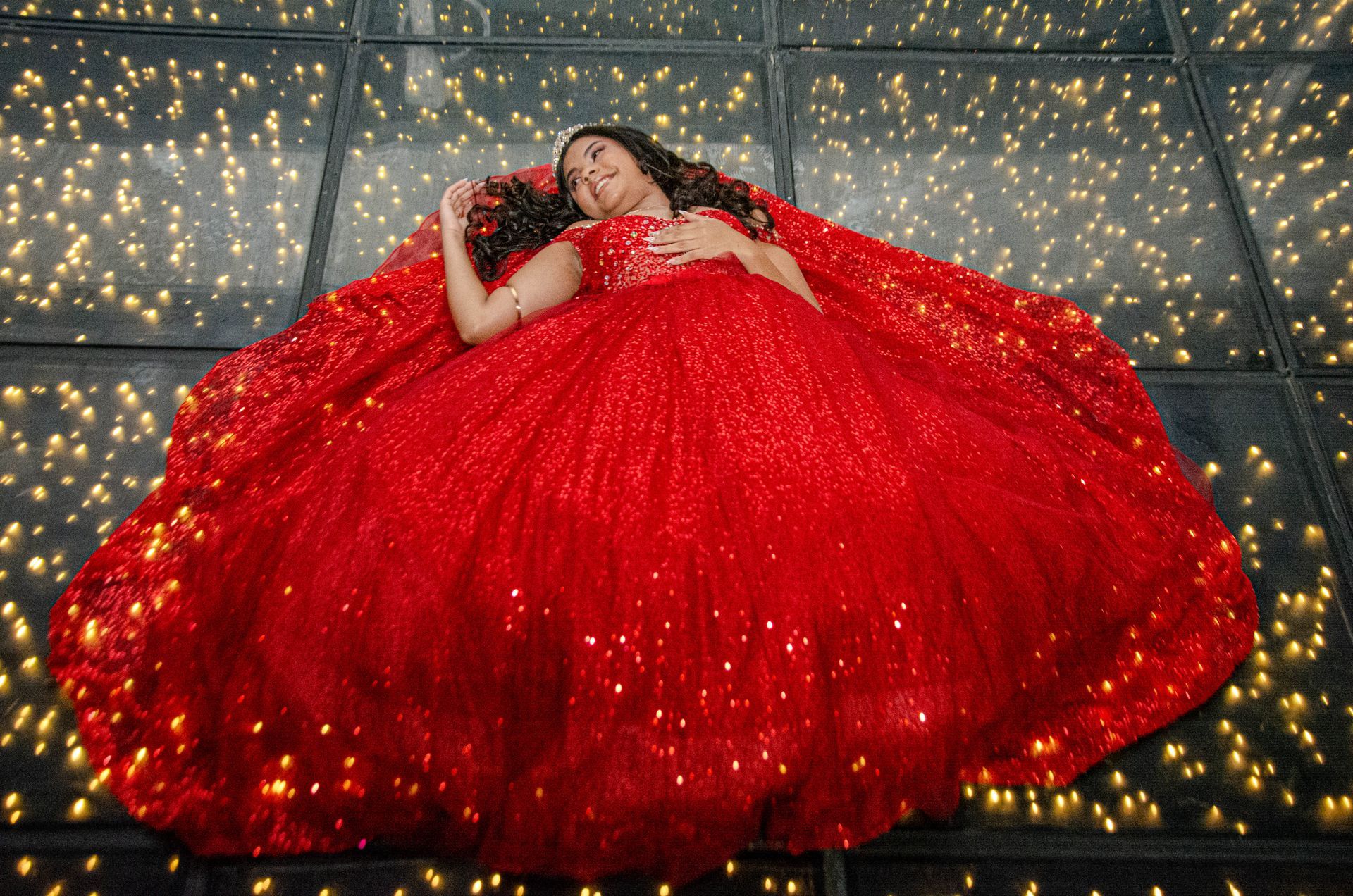 Mulher em vestido vermelho de lantejoulas está deitada no chão brilhante, com a cabeça apoiada e os braços ao lado do corpo.