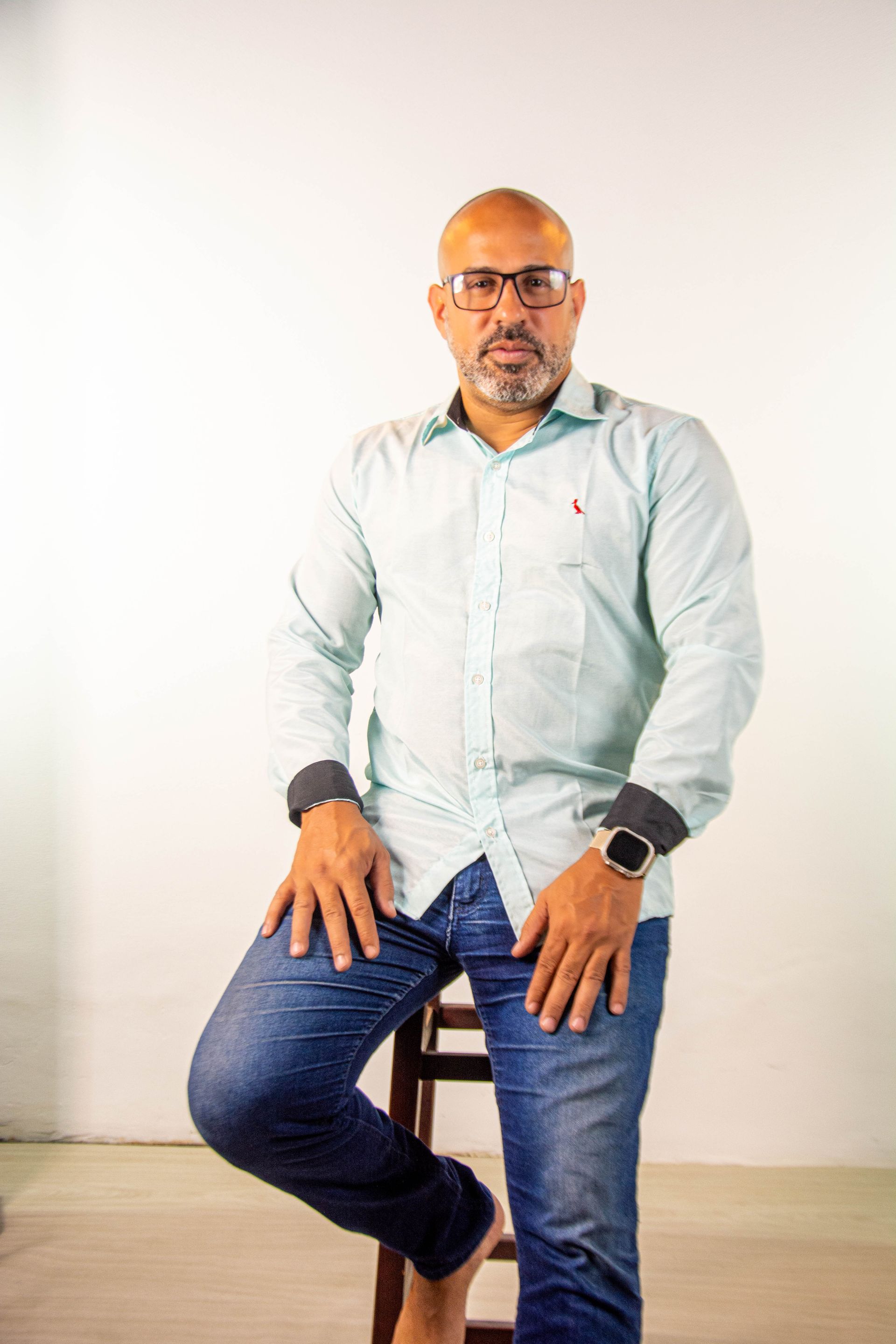 Homem de camisa azul e jeans sentado em um banquinho, olhando para a câmera.