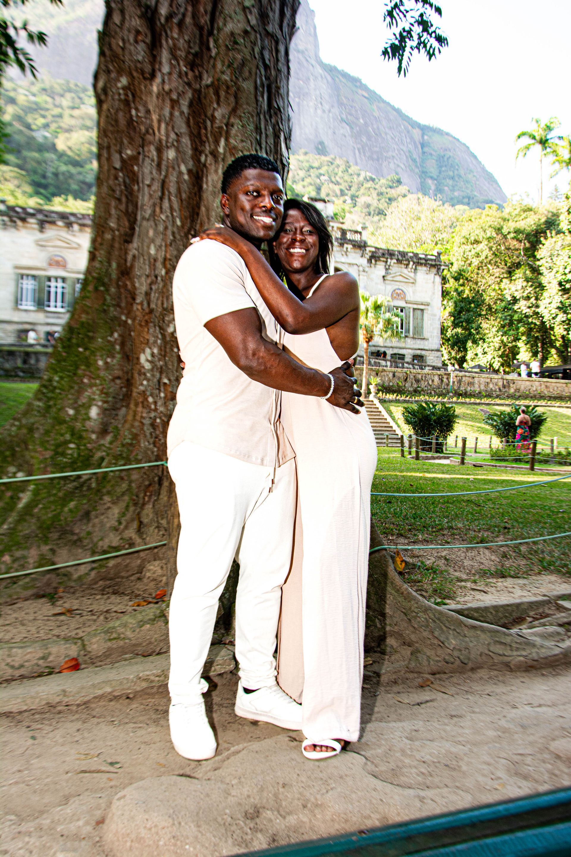 Casal se abraçando, sorrindo ao ar livre; árvore, prédio, montanha ao fundo.