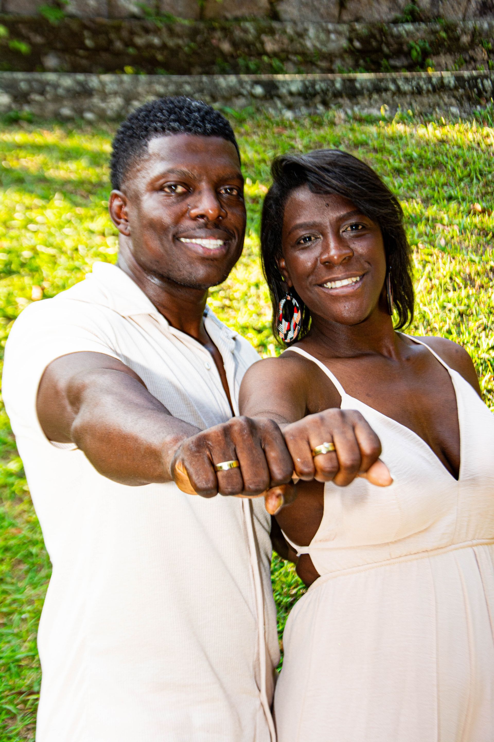 Casal sorrindo, exibindo alianças de ouro. Fundo claro, ambiente externo.