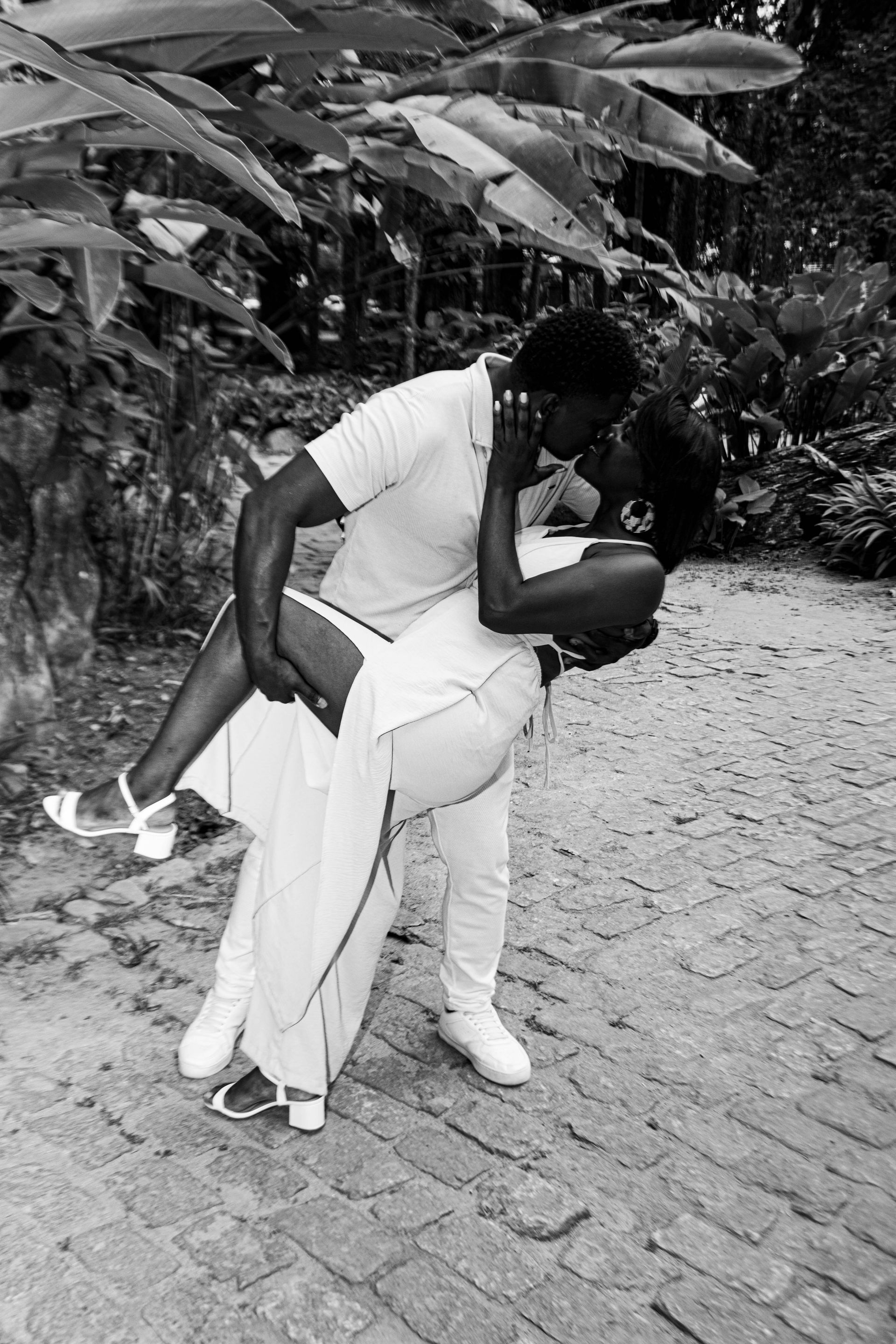 Casal se beijando enquanto dança ao ar livre, homem segurando mulher, ambos em roupas brancas.