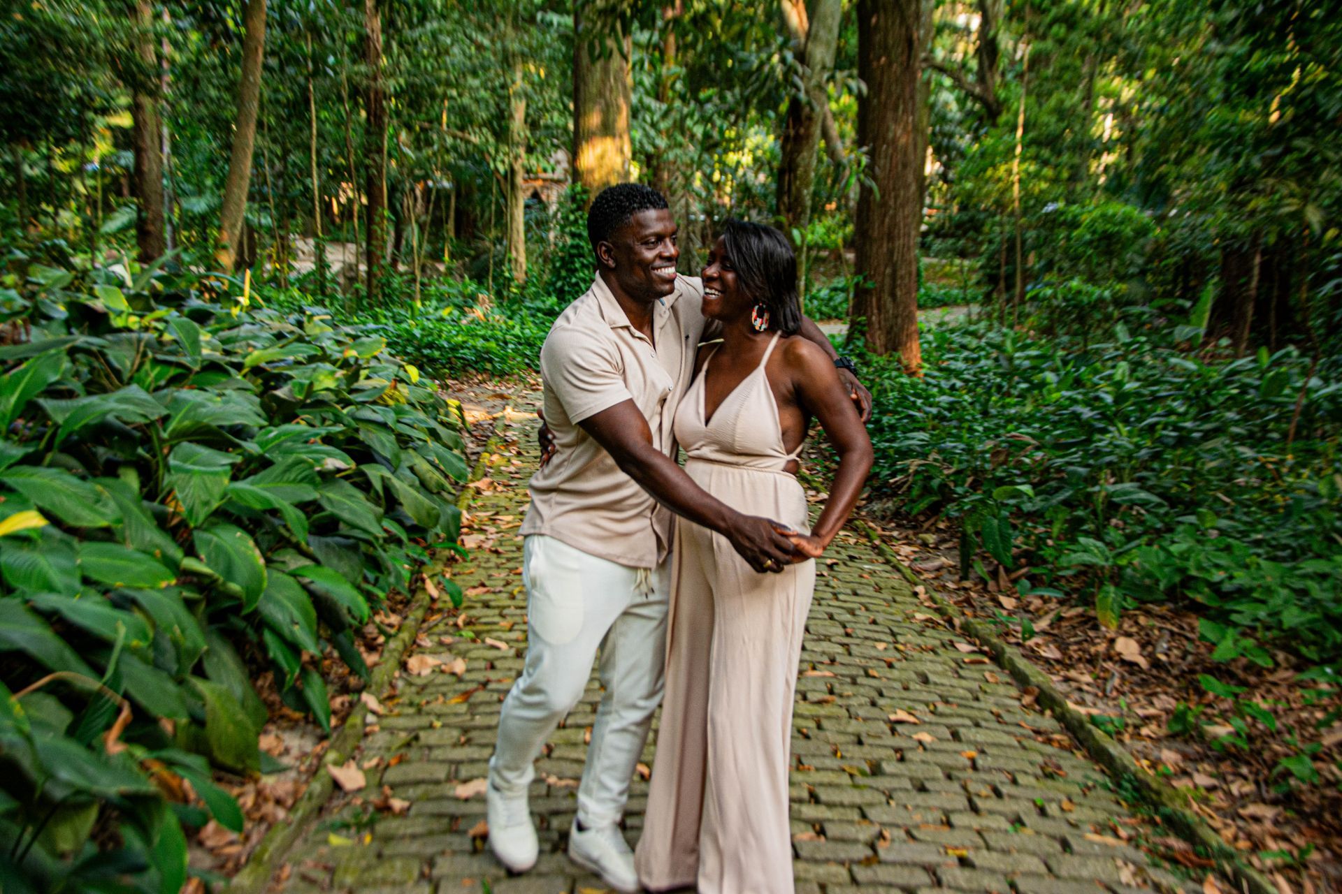 Casal se abraça em uma trilha de tijolos em um parque verdejante. A mulher usa um vestido leve, o homem, uma camisa bege e calça branca.