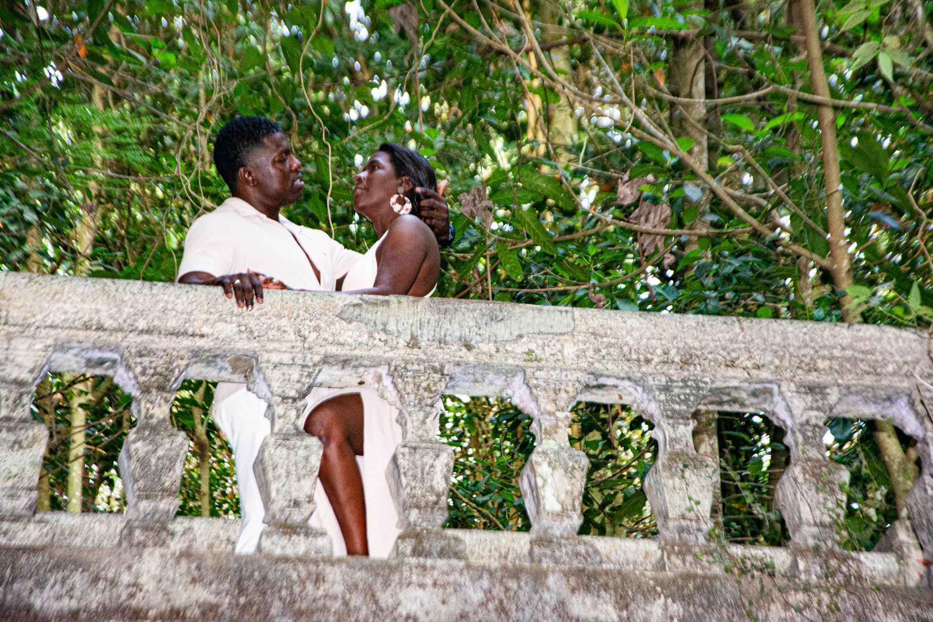 Casal se olhando por cima de um corrimão de pedra desgastado. Folhagem verde ao fundo.