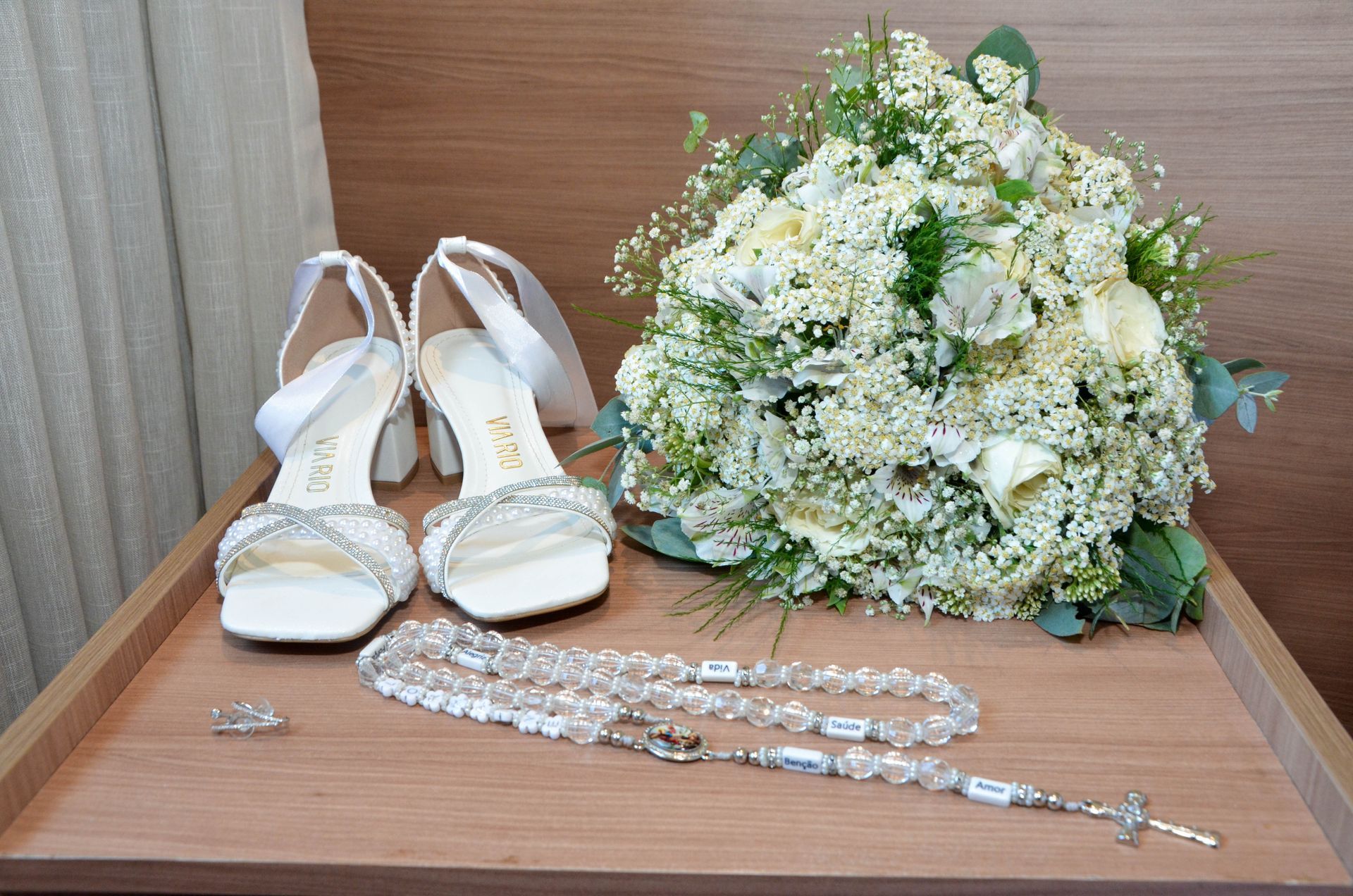 Sapatos de casamento, buquê, rosário e anel em uma superfície de madeira.