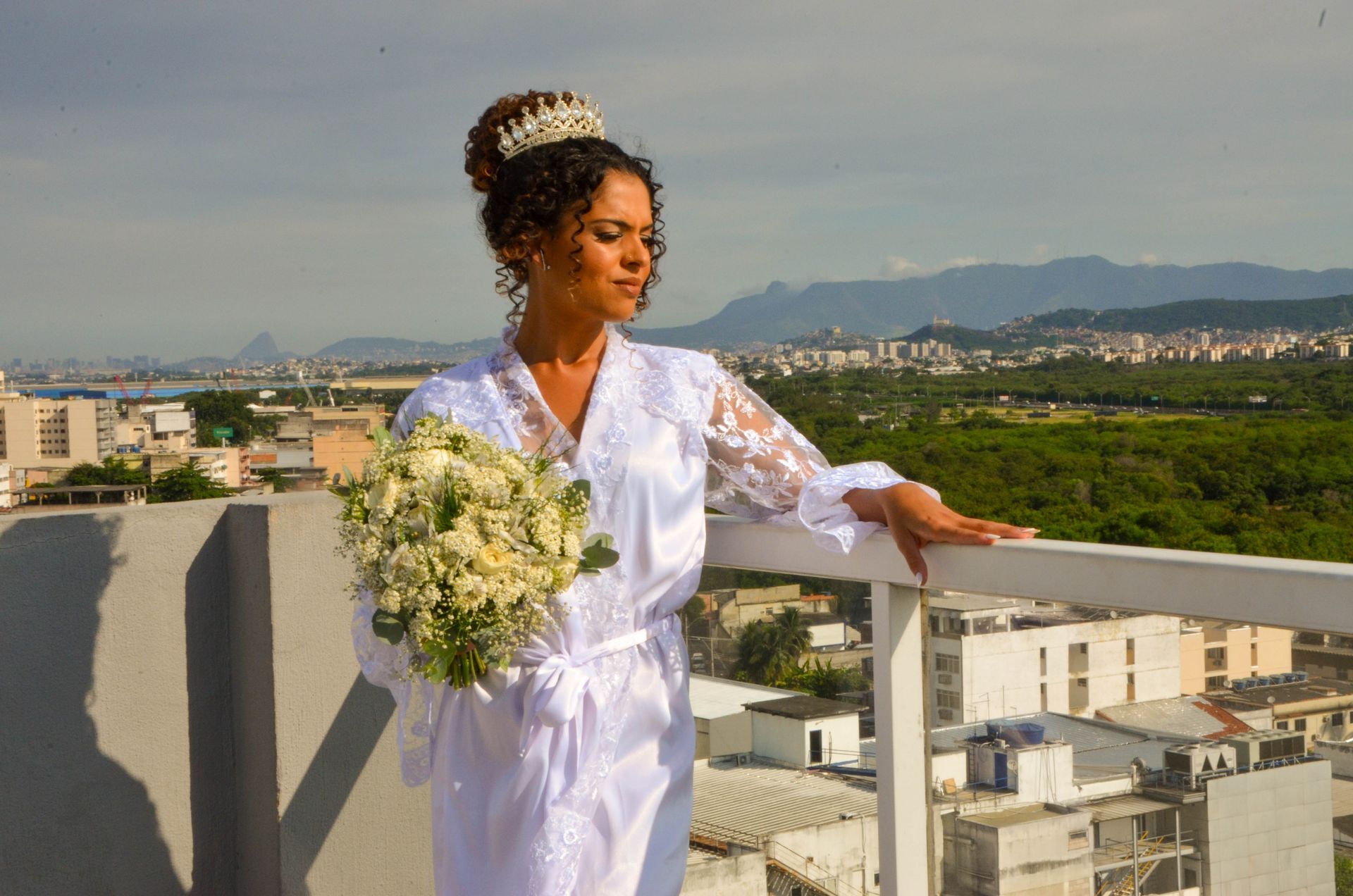 Noiva de túnica branca e coroa segurando buquê, observando a cidade do alto de um telhado.