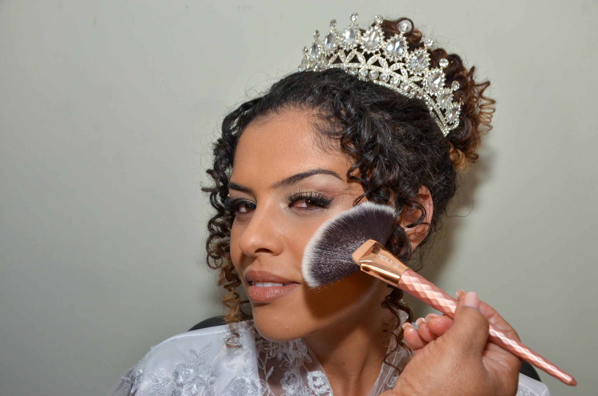 Pessoa se maquiando, usando uma tiara e segurando um pincel de blush.