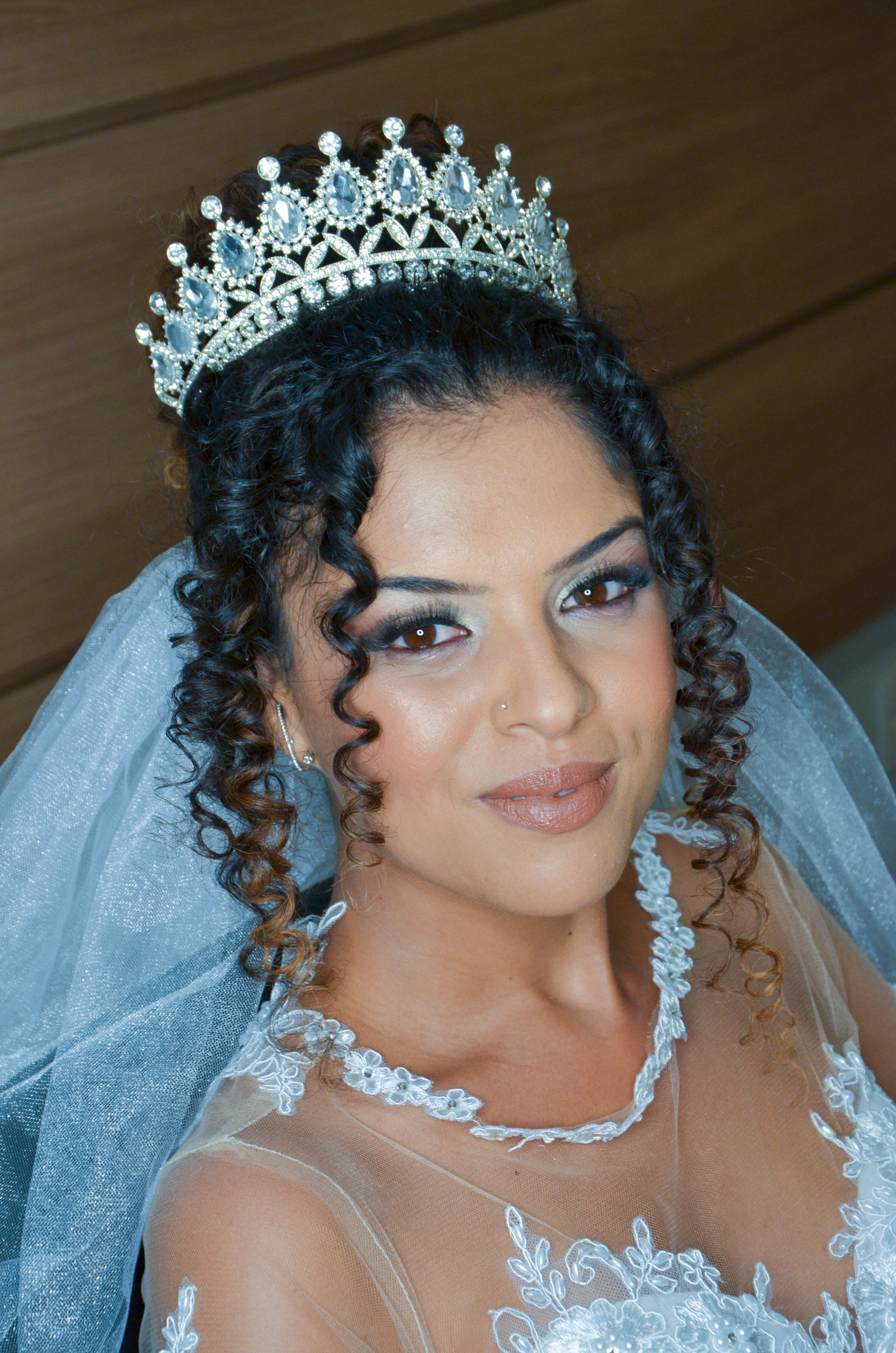 Noiva usando tiara e véu cravejados de joias, sorrindo. Ela usa um vestido branco e tem cabelos castanhos cacheados.