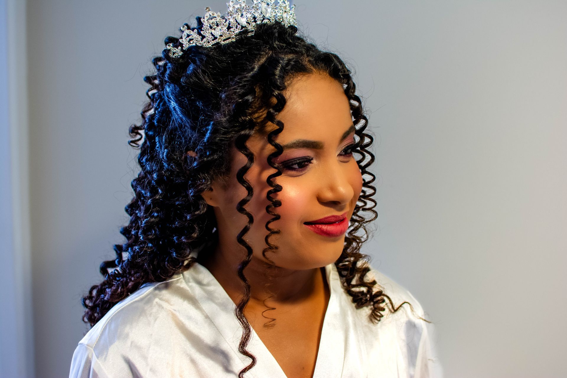 Mulher com cabelos cacheados e tiara, vestindo um robe branco, com batom vermelho e sombra rosa.