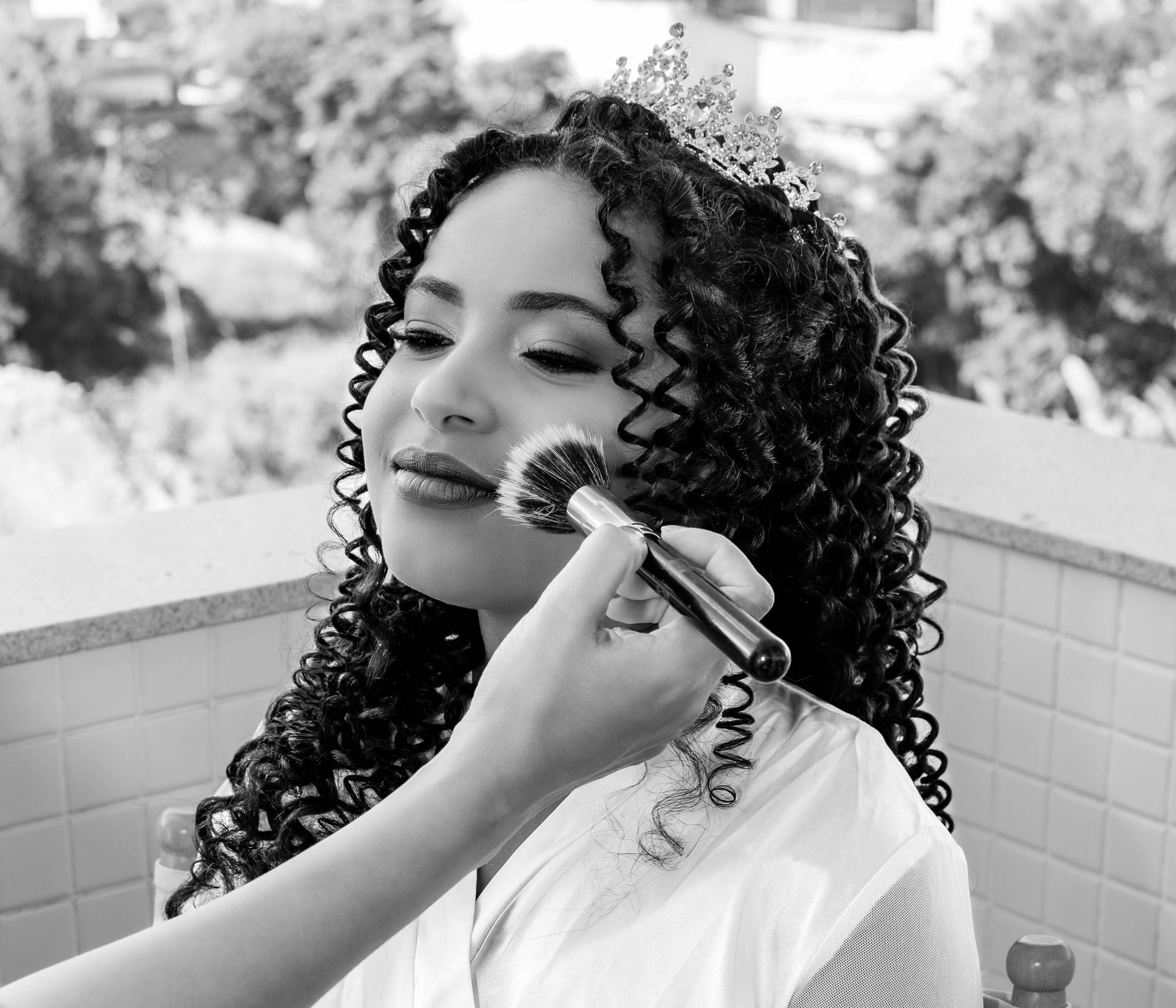 Pessoa recebendo aplicação de maquiagem; tem cabelo cacheado, tiara, ambiente externo.