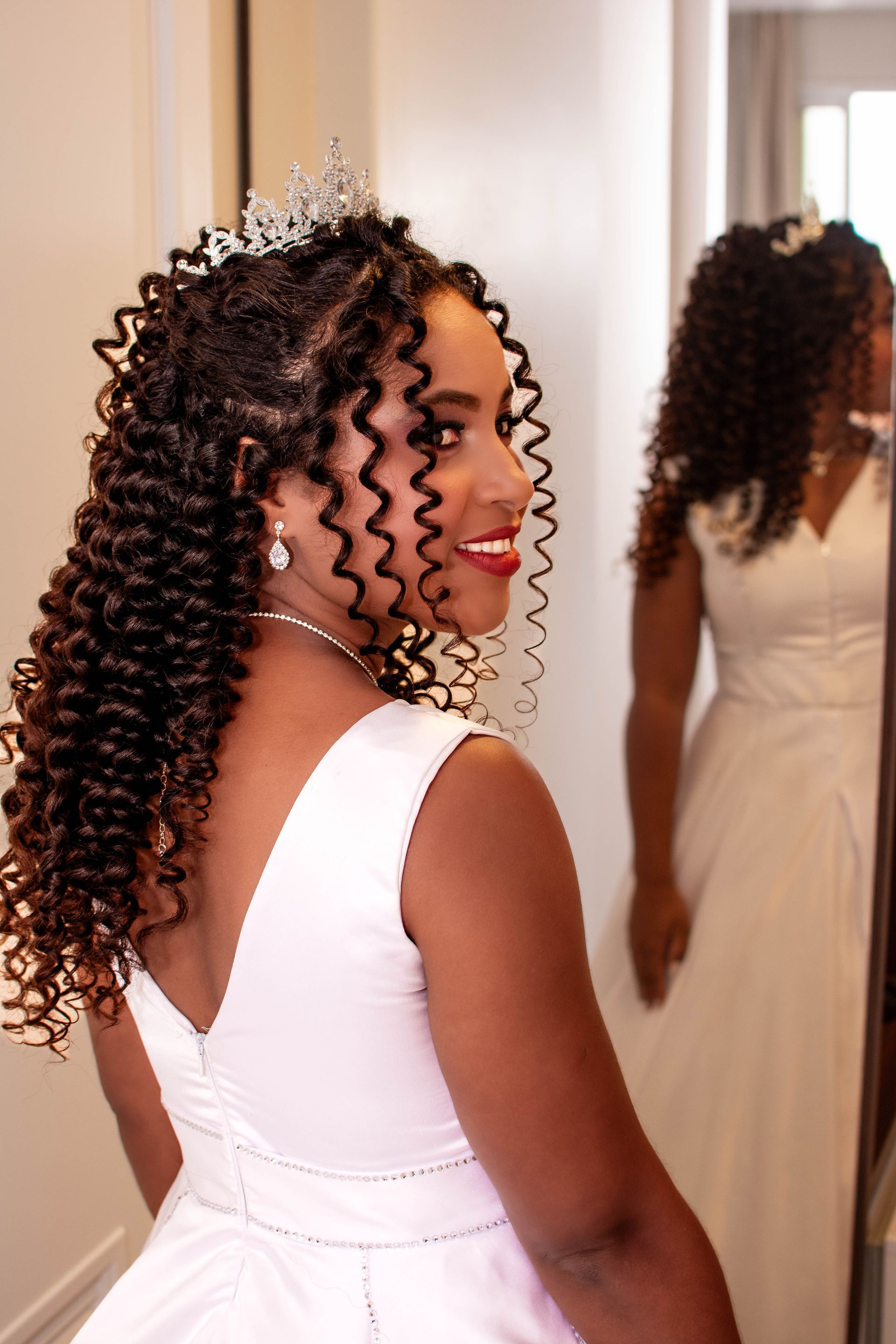 Mulher de vestido branco, tiara e cabelo cacheado sorri para seu reflexo no espelho.
