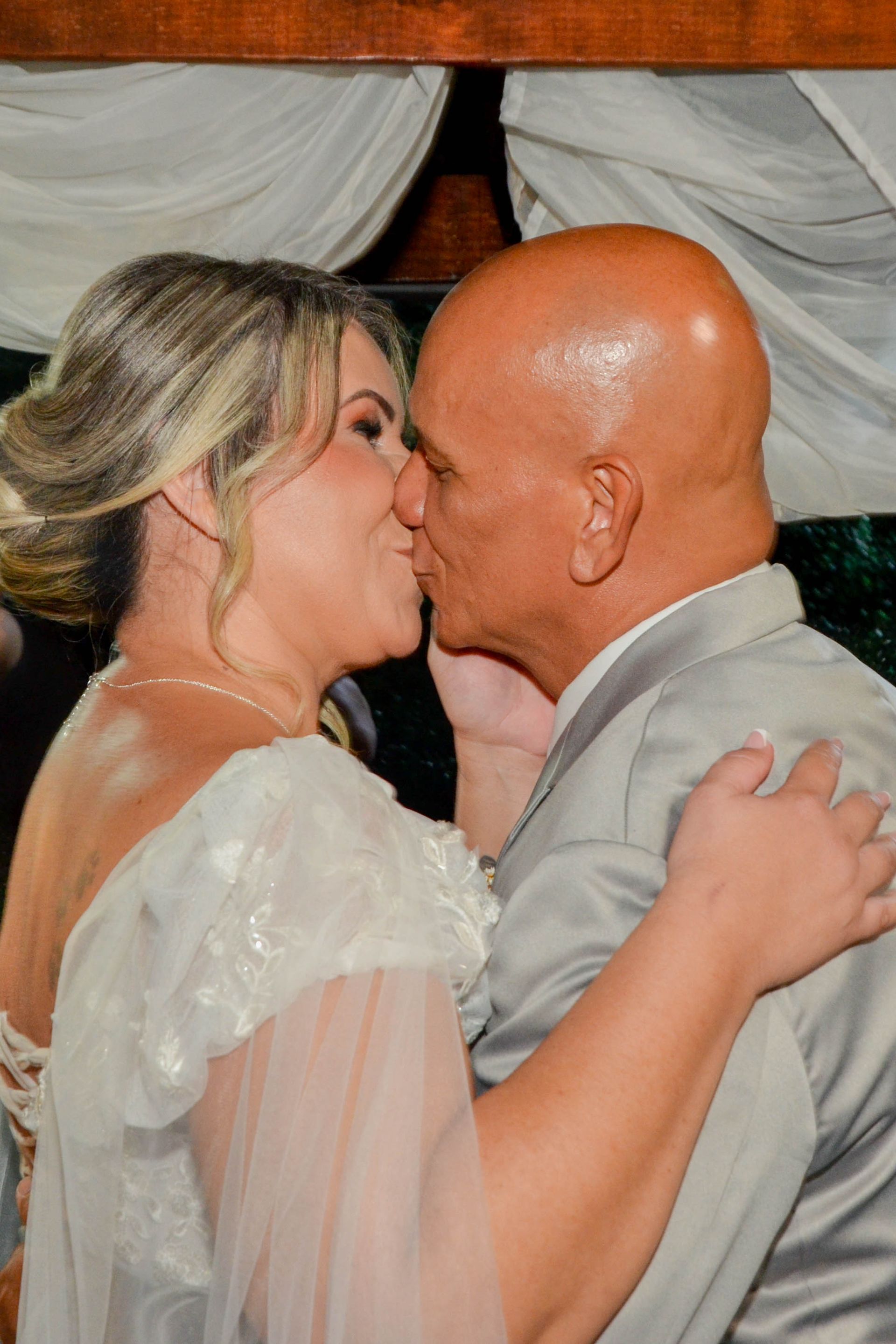 Noiva de vestido branco beijando um homem de terno cinza. Eles se abraçam em um ambiente externo.