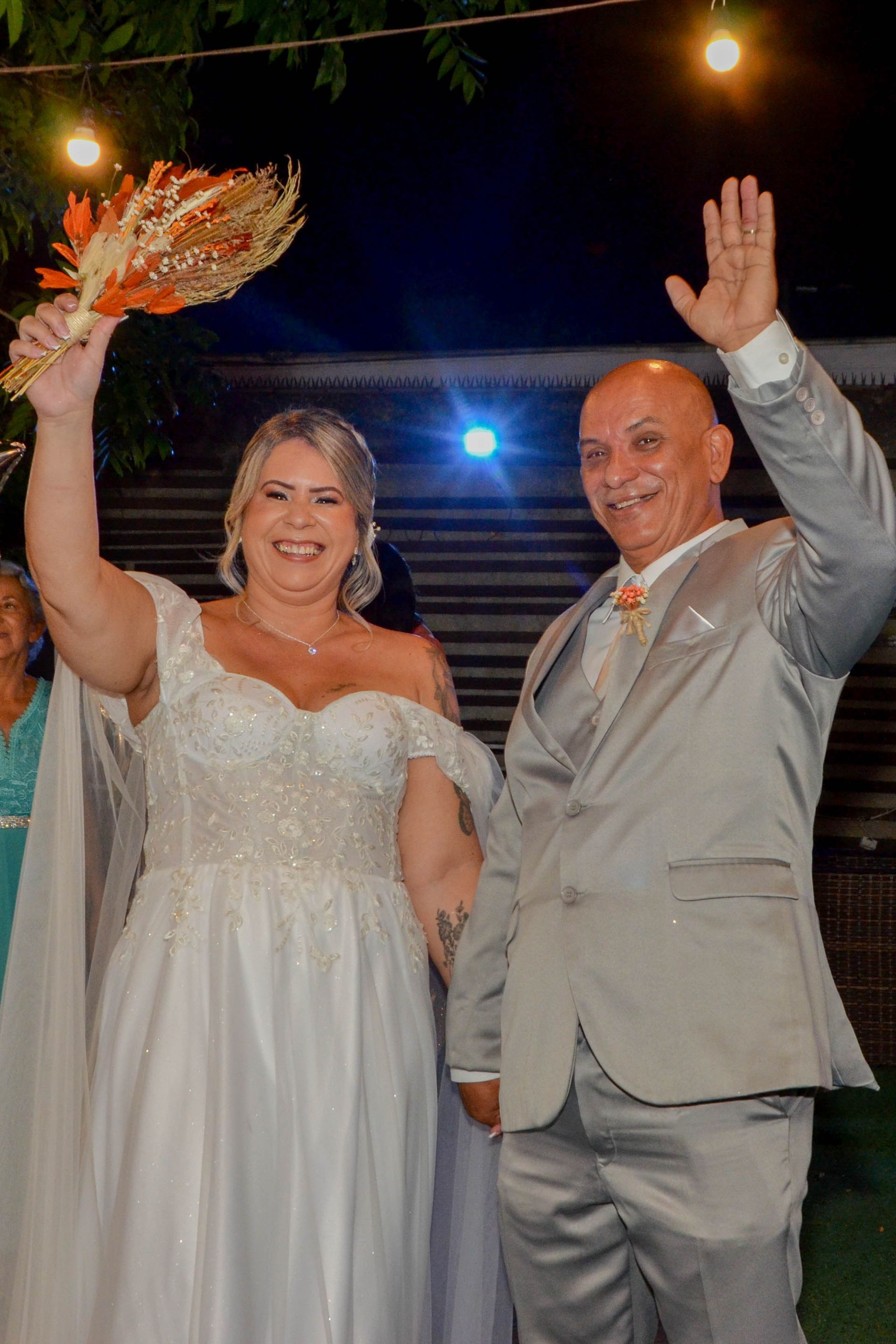 Recém-casados acenando, noiva de vestido branco segurando buquê, noivo de terno cinza, em um evento noturno ao ar livre.