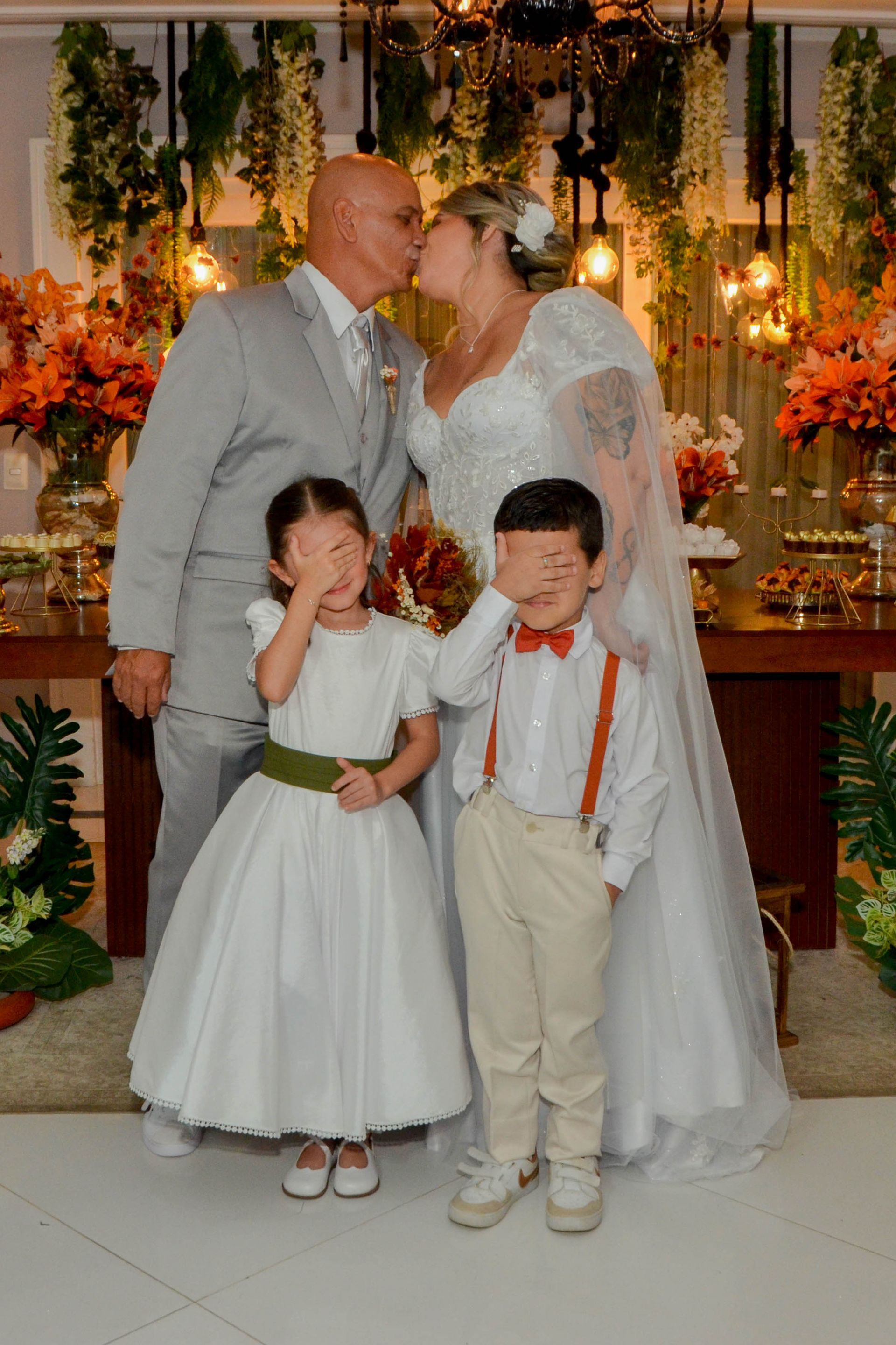Recém-casados se beijando, ladeados por duas crianças cobrindo os olhos, em uma recepção de casamento com decoração floral.