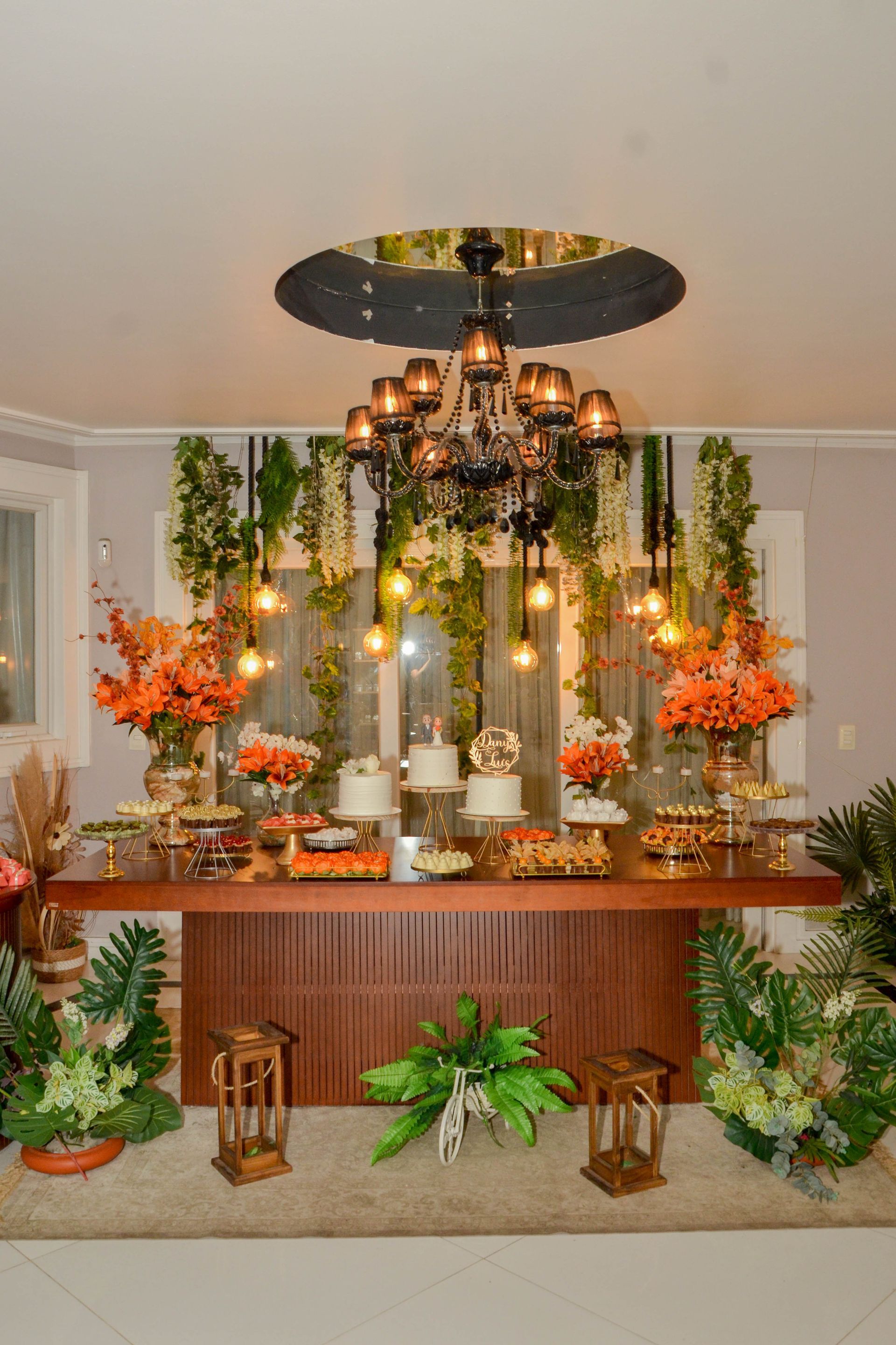 Mesa de sobremesas com bolos, flores e luzes penduradas; cercada por vegetação e detalhes em madeira.