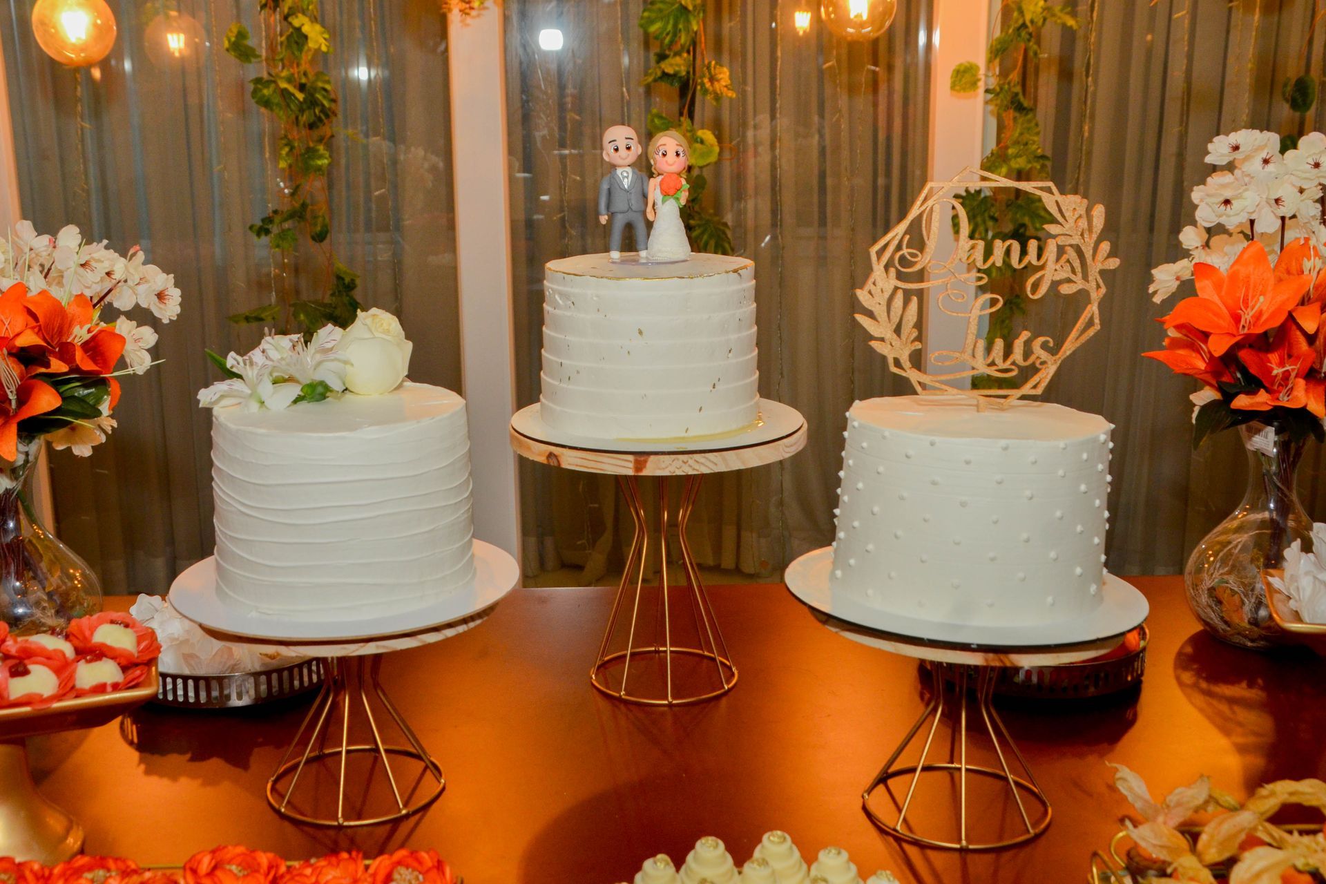 Três bolos brancos em suportes dourados, decorados para uma celebração. Flores de laranjeira e outras guloseimas os cercam.