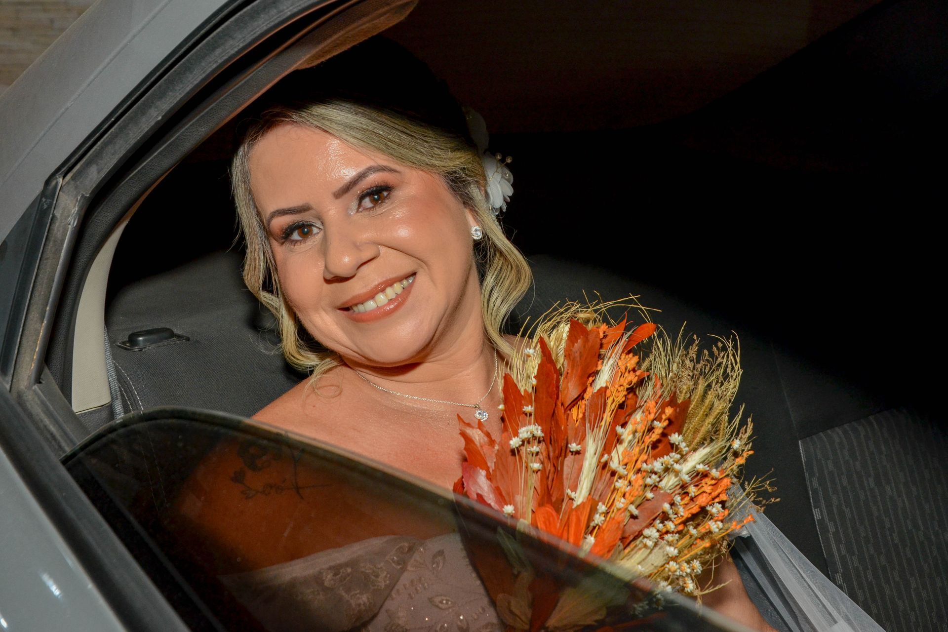 Noiva em um carro, sorrindo, segurando um buquê de laranjas. Ela usa um vestido branco e tem cabelos loiros.