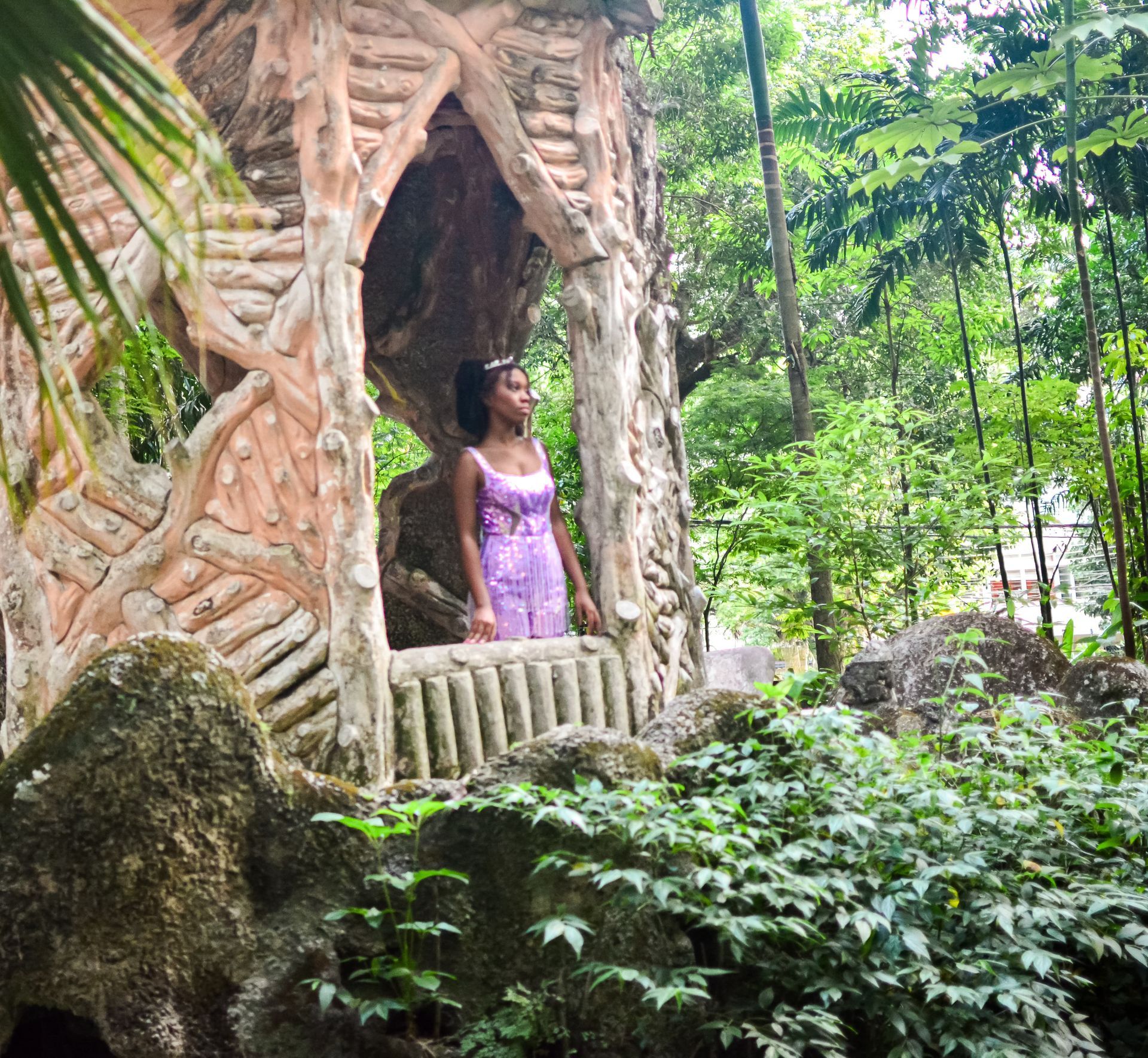 Mulher de vestido roxo observa de uma estrutura de pedra em um cenário de floresta verdejante.
