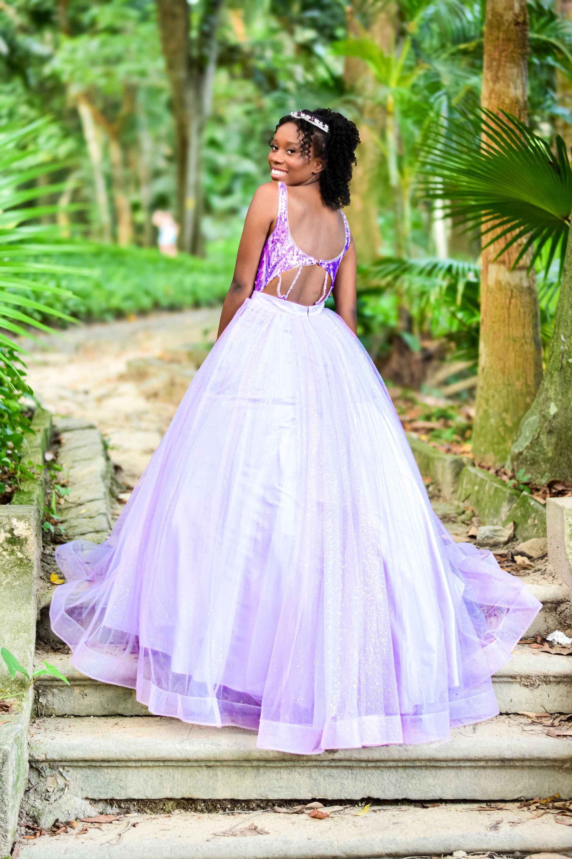 Mulher em um vestido formal lilás sorri por cima do ombro em degraus de pedra ao ar livre com vegetação exuberante ao fundo.