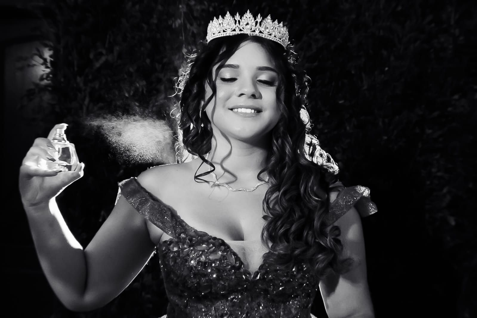 Mulher em traje formal borrifa perfume e usa uma coroa.