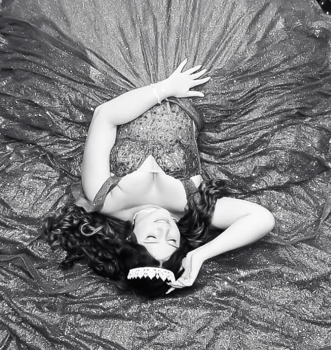 Mulher com vestido brilhante, deitada, sorrindo. Ela usa uma coroa e uma mão perto do rosto.