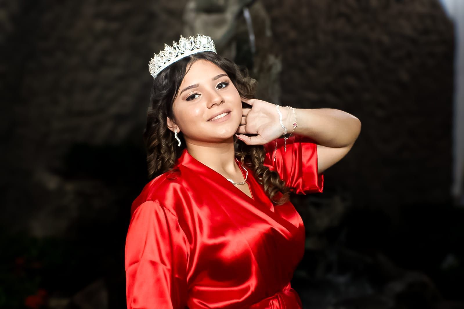 Mulher usando uma coroa e um manto vermelho sorri, posando perto de um fundo escuro.