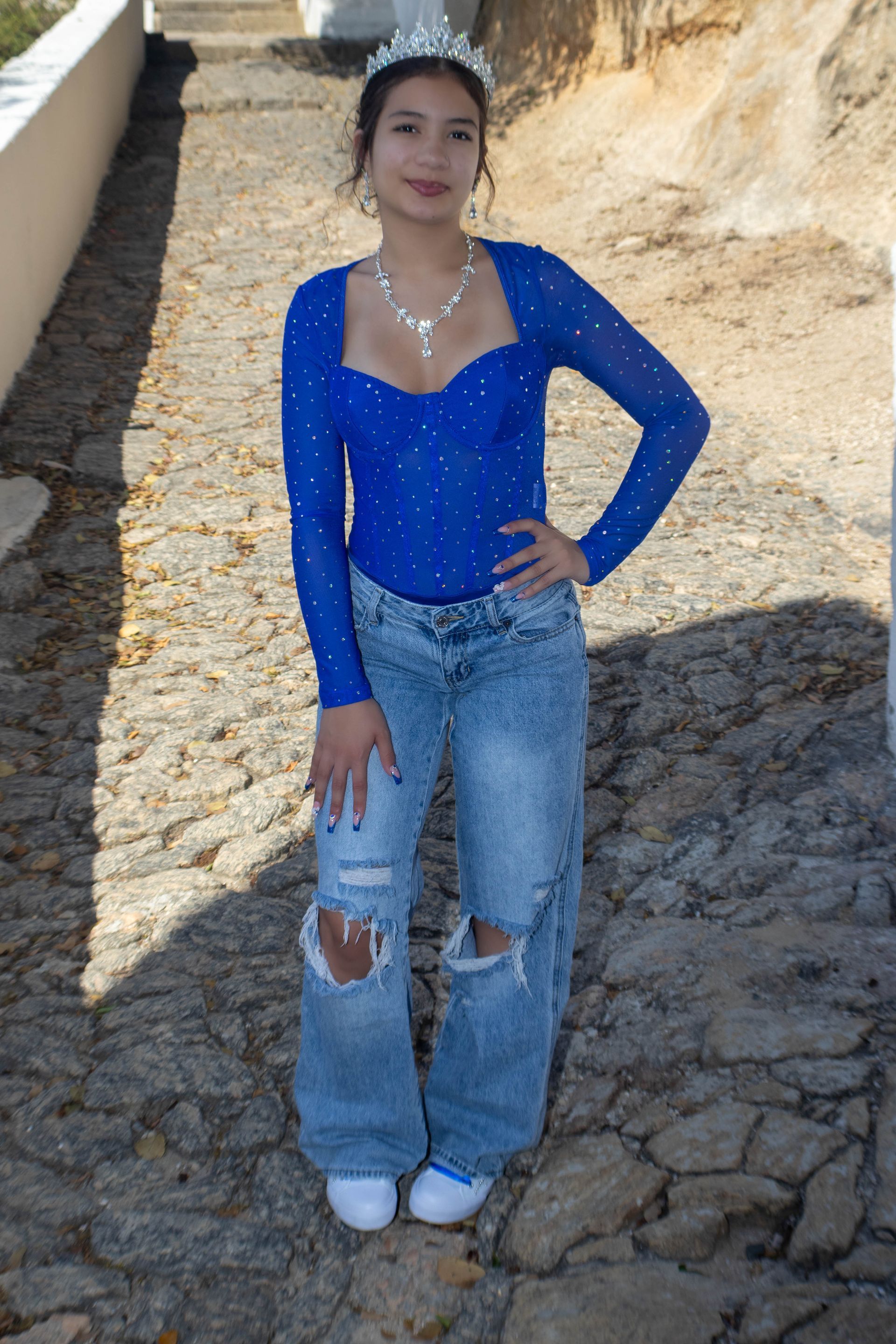 Mulher de blusa azul e jeans rasgados, usando uma tiara, posa em um caminho de pedra.