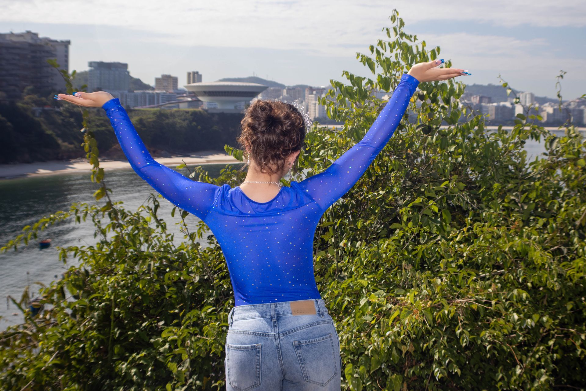Mulher com os braços levantados, blusa azul e jeans, com vista para uma baía e cidade ao fundo.