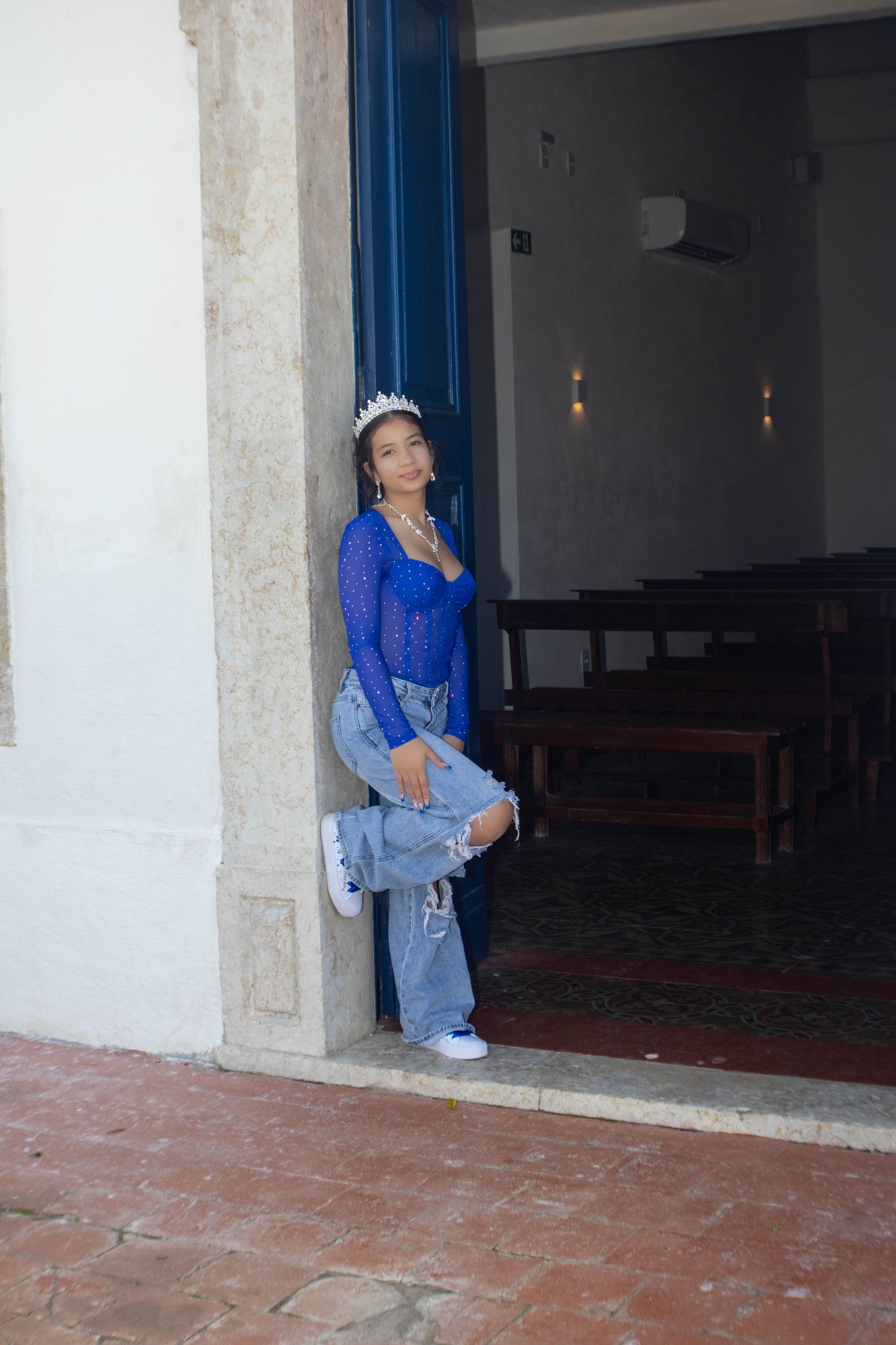 Mulher de blusa azul e calça jeans rasgada encostada no batente de uma porta azul, com tênis brancos. Cenário de igreja.