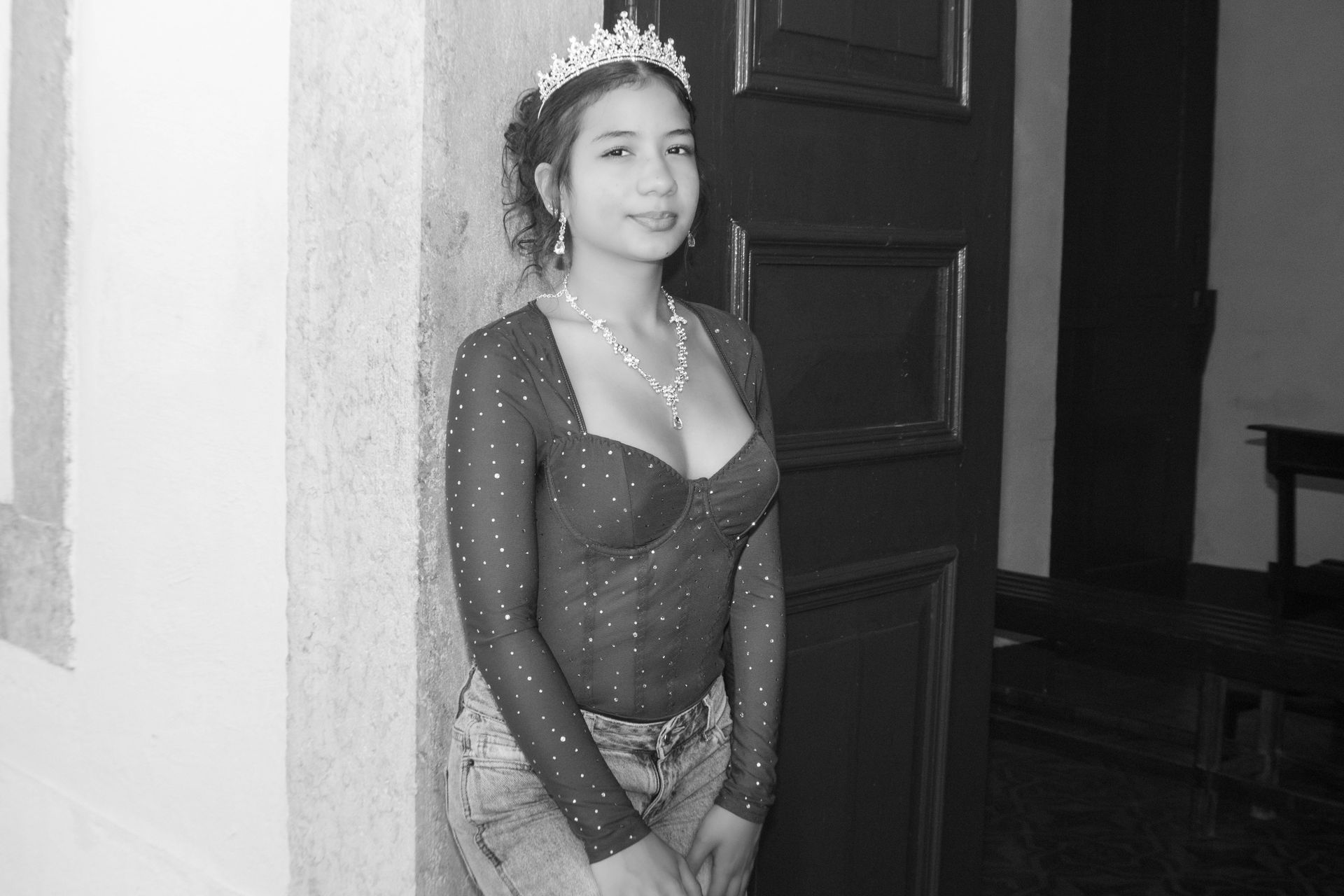Mulher usando uma coroa, blusa transparente e jeans está em pé perto de uma porta de madeira escura.