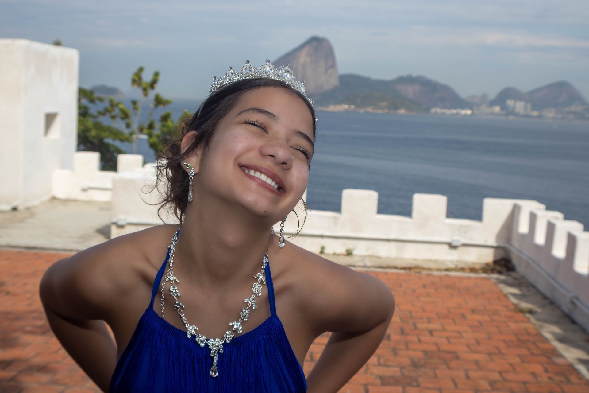 Mulher usando uma tiara e joias sorri em frente a uma paisagem costeira com montanhas.