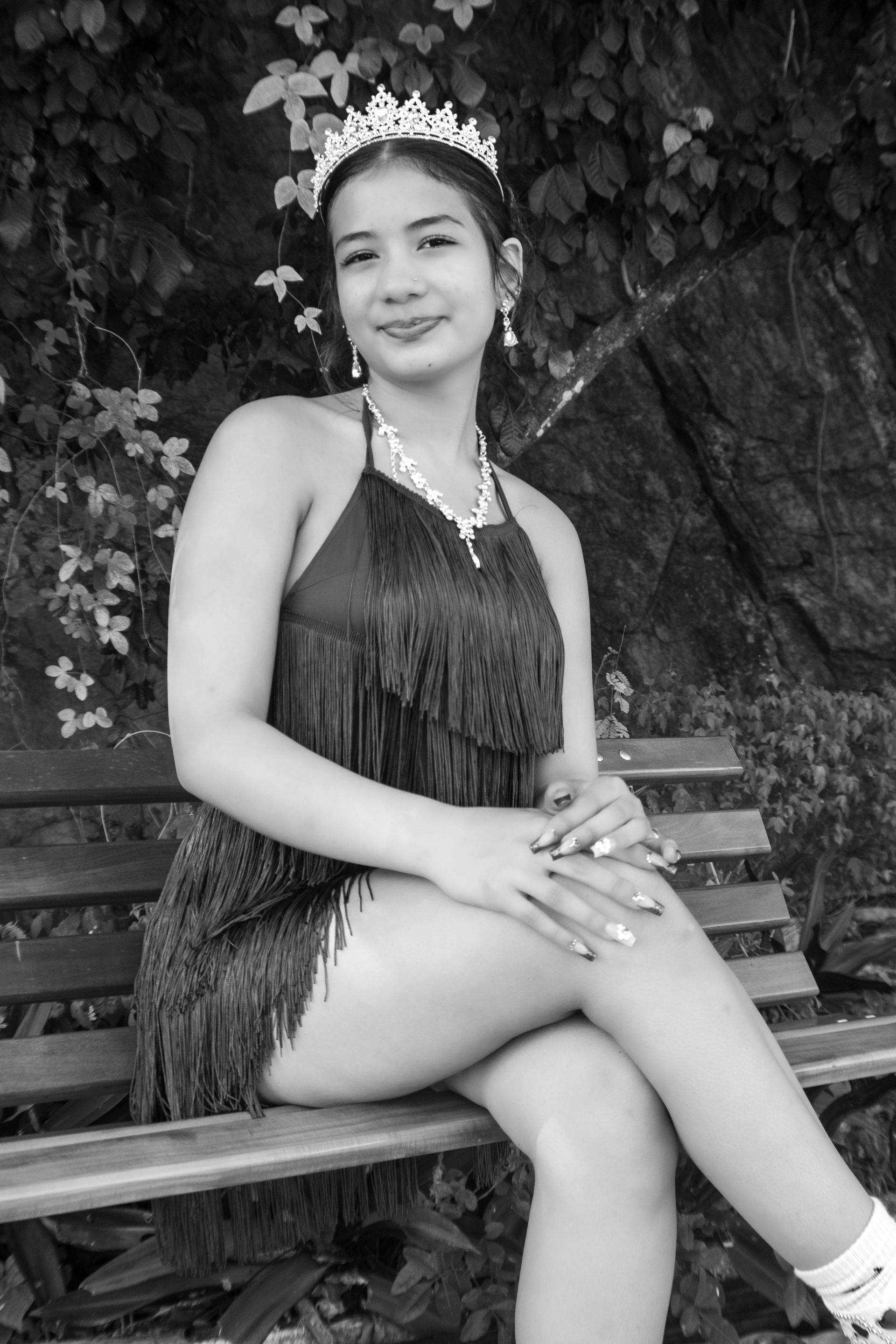 Mulher com vestido franjado e tiara sentada em um banco, sorrindo. Ambiente externo com folhagens.