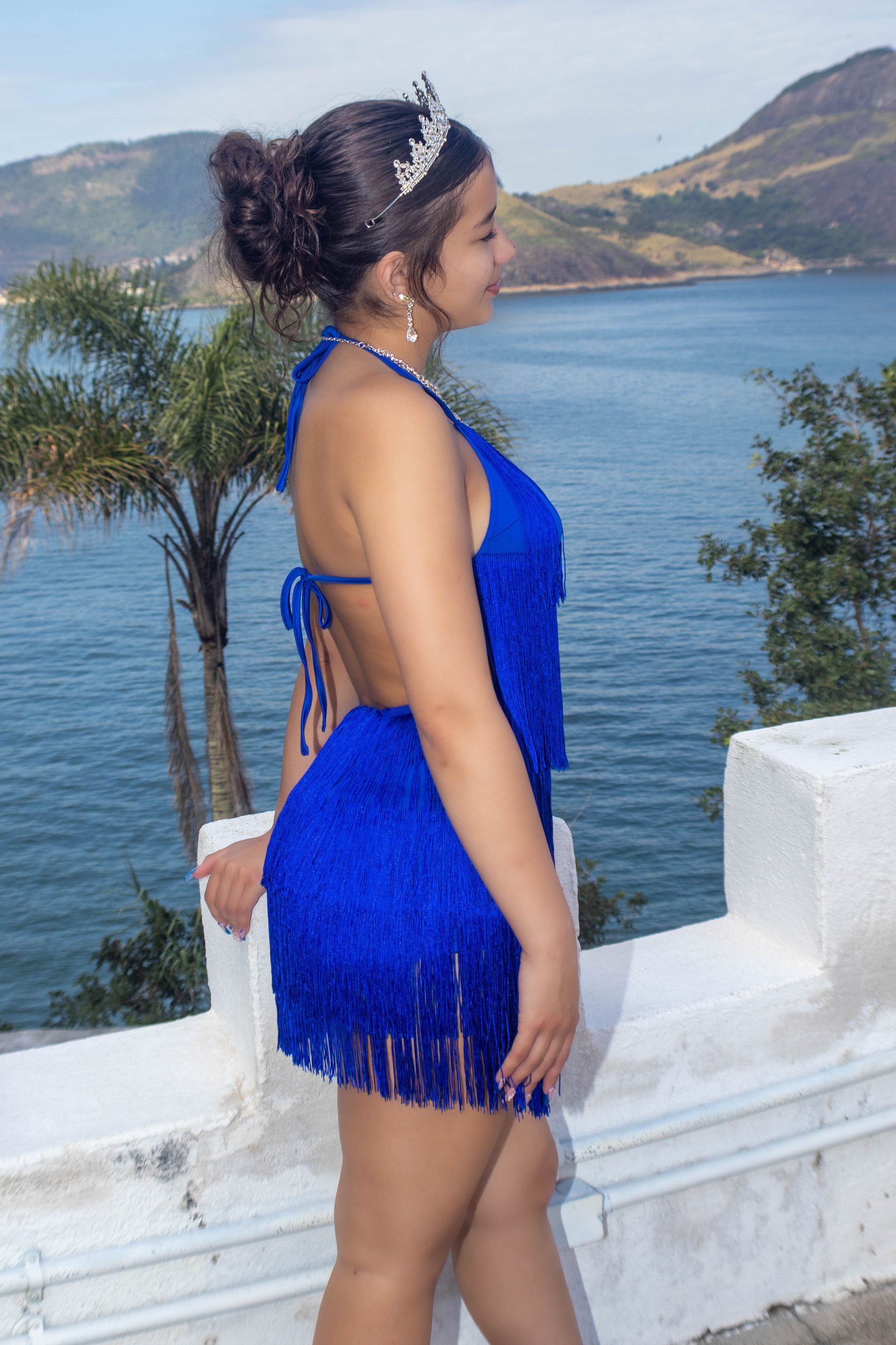 Mulher com vestido azul com franjas e tiara posa em frente a uma vista costeira.
