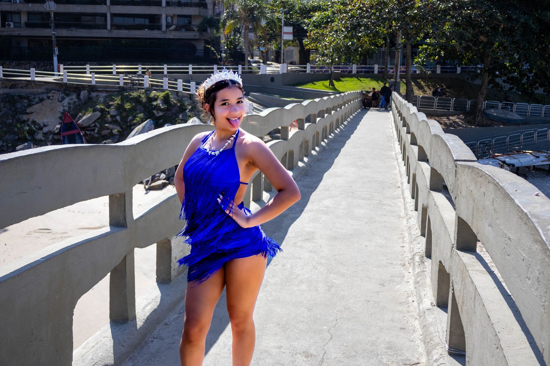 Mulher de vestido azul com a língua para fora, em pé em uma ponte perto de uma praia.