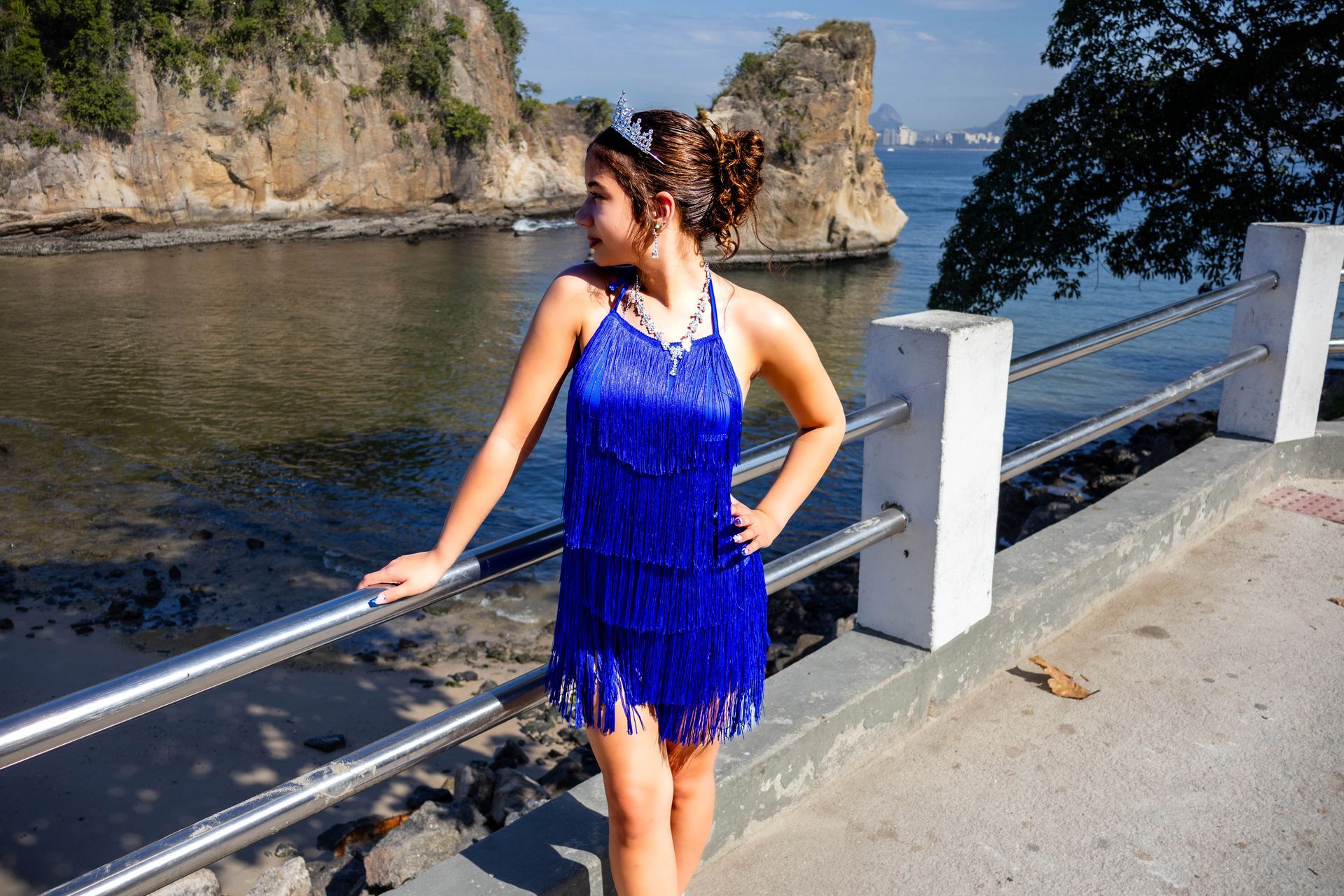 Mulher com vestido azul com franjas, posando perto de uma grade perto da água com penhascos rochosos ao fundo.