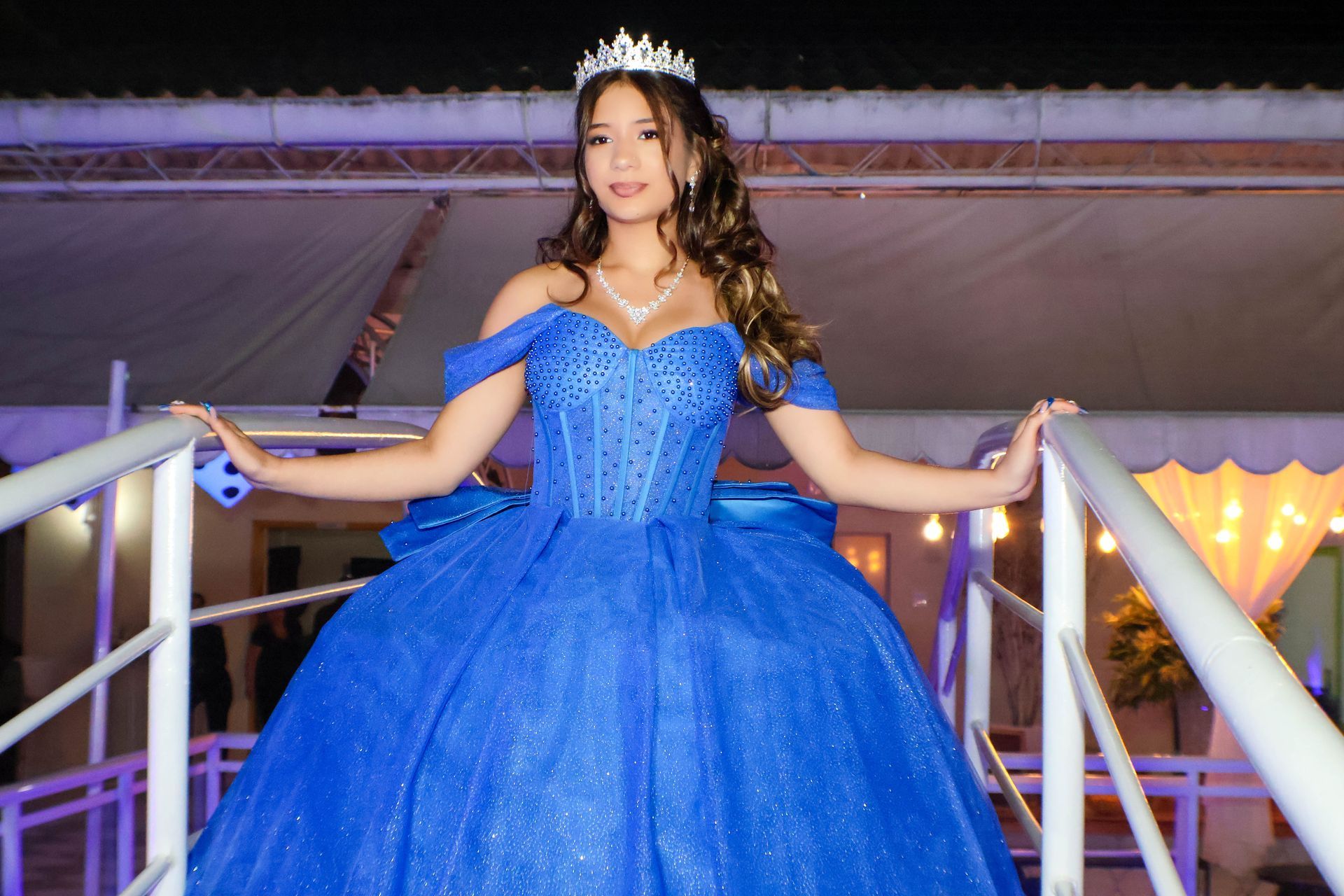 Mulher de vestido azul e tiara posa em uma ponte branca com um cenário de festa ao fundo.