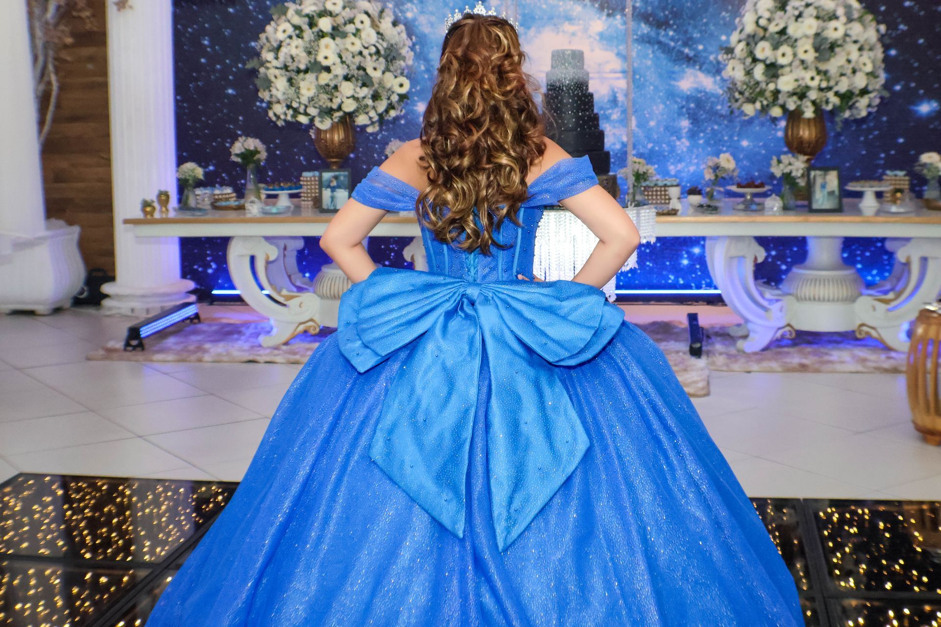 Mulher em vestido de baile azul com laço grande, posando em uma festa com decoração floral.