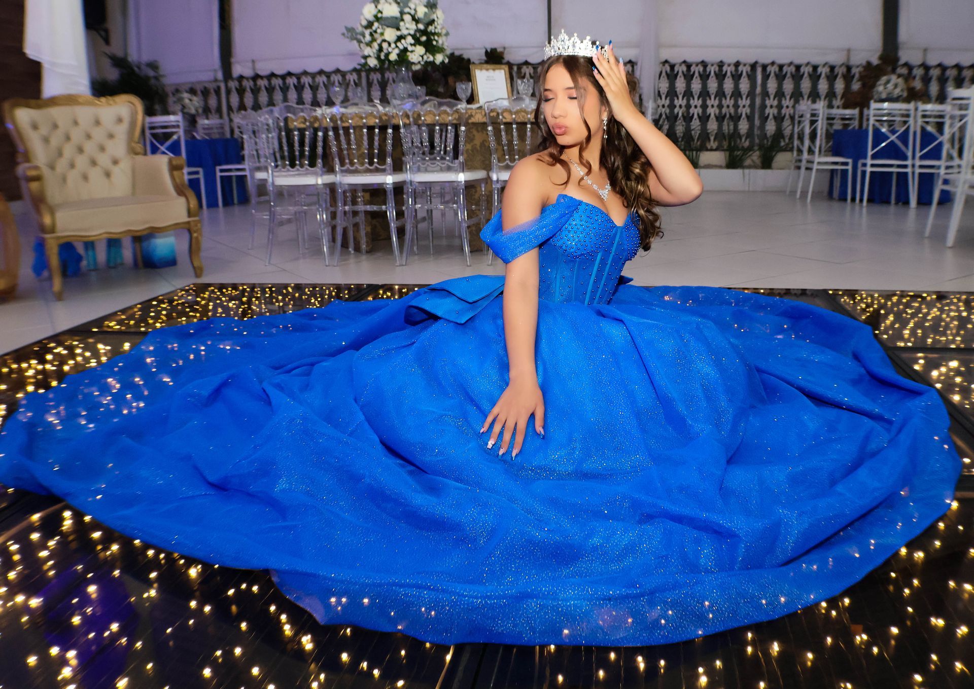 Mulher com um vestido azul royal com uma coroa, sentada em uma pista de dança com um padrão iluminado, em uma sala decorada.