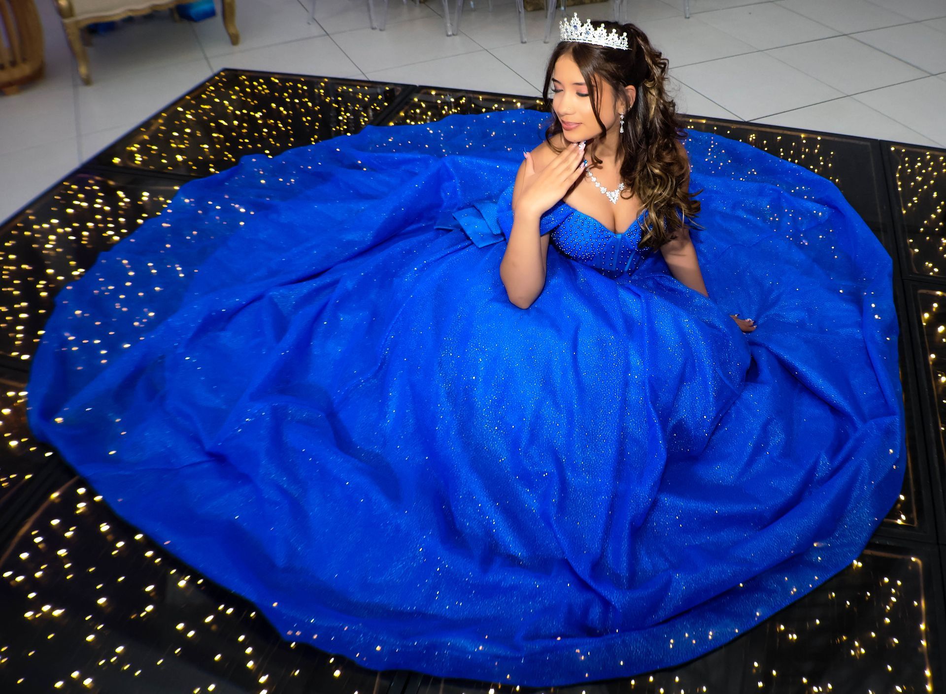 Mulher com um vestido de baile azul brilhante com uma tiara, sentada no palco, olhando para baixo.