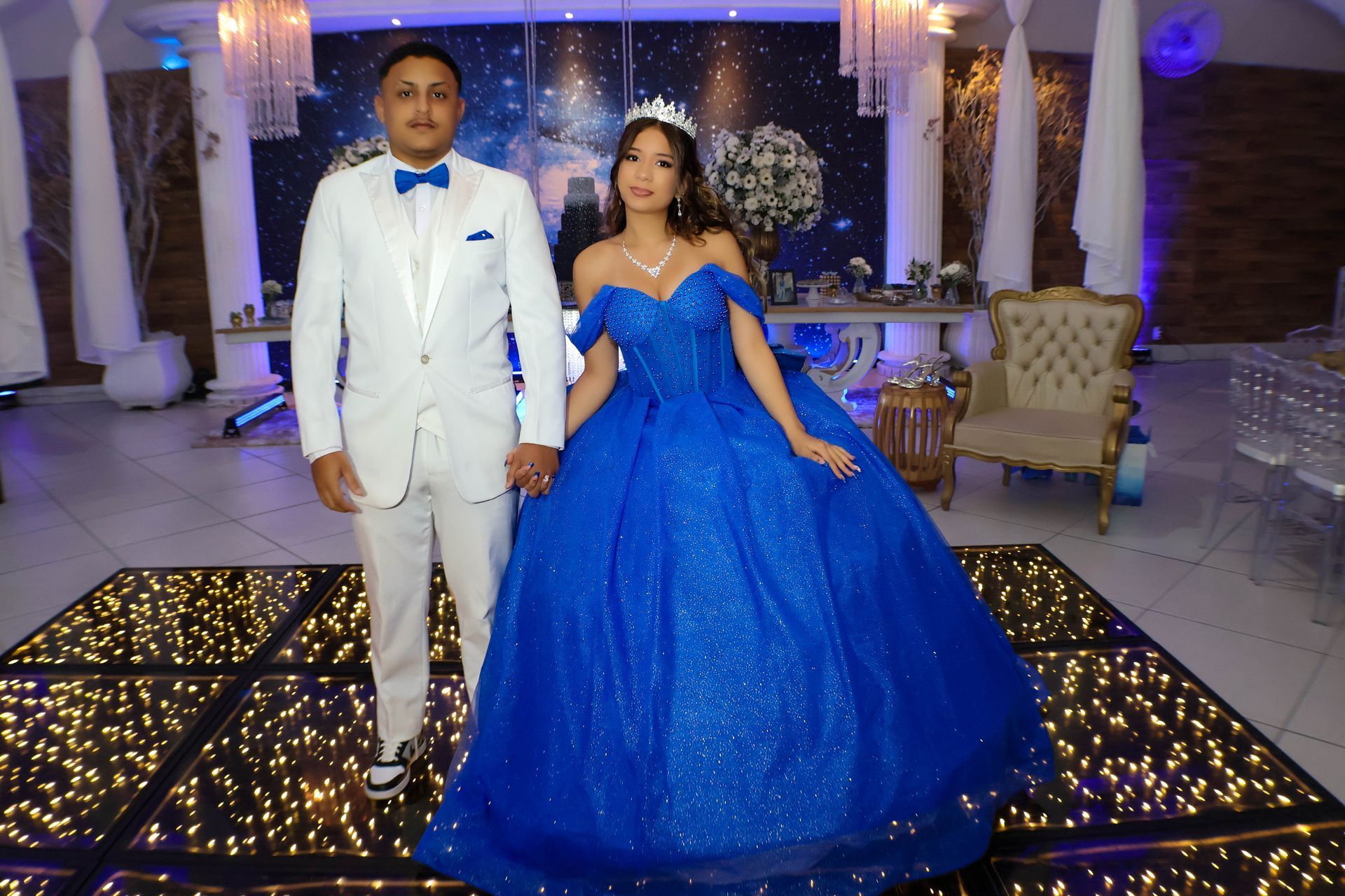 Casal, homem de terno branco e mulher de vestido azul, de mãos dadas em uma pista de dança iluminada. Fundo festivo.