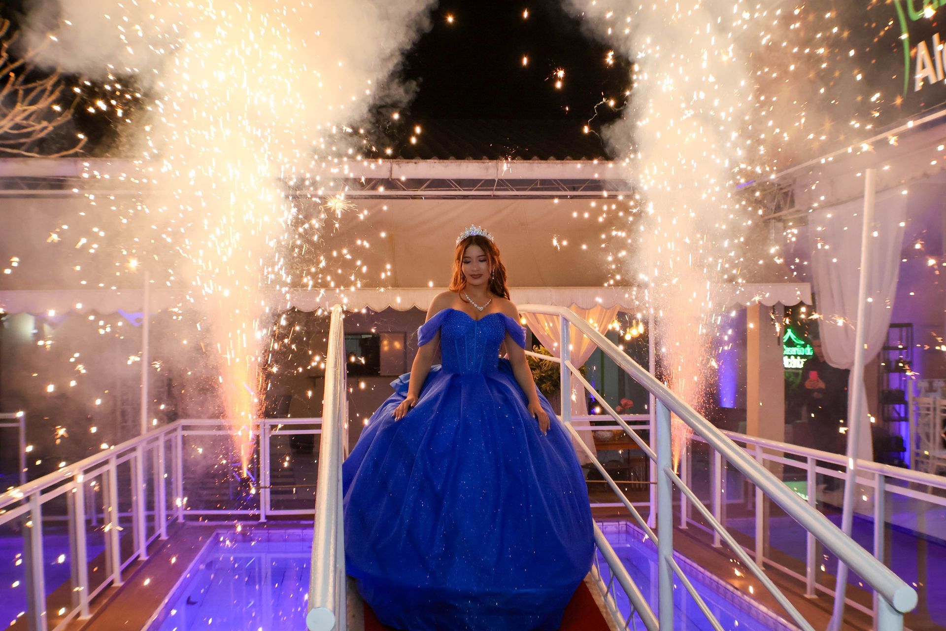 Mulher de vestido azul desce uma rampa com fogos de artifício, em uma celebração noturna.